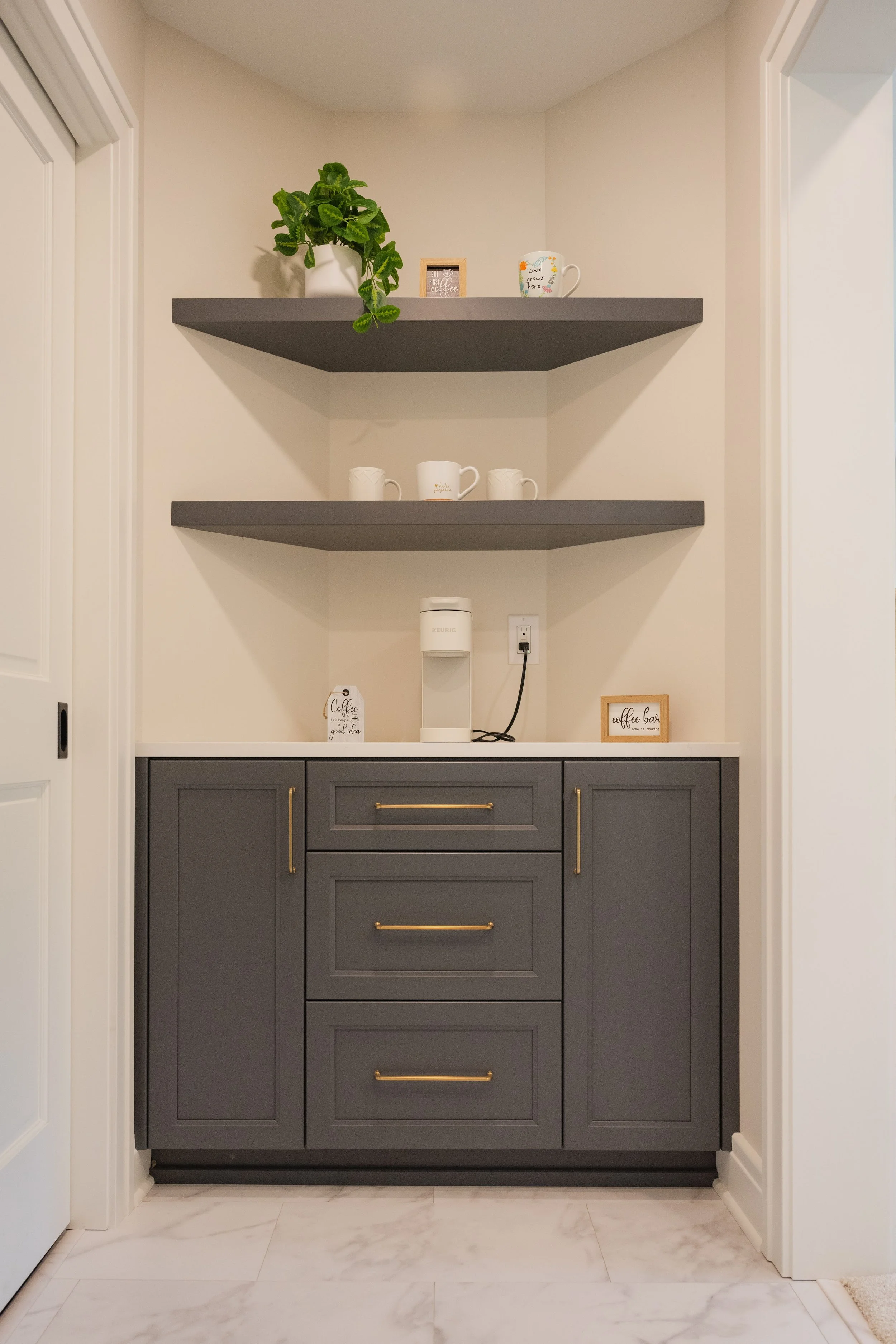 Dark gray cabinet with gold handles in a corner with two gray floating shelves above it. The shelves hold a potted plant, small framed picture, cup, and other decor items. The space has a marble tile floor and white walls.