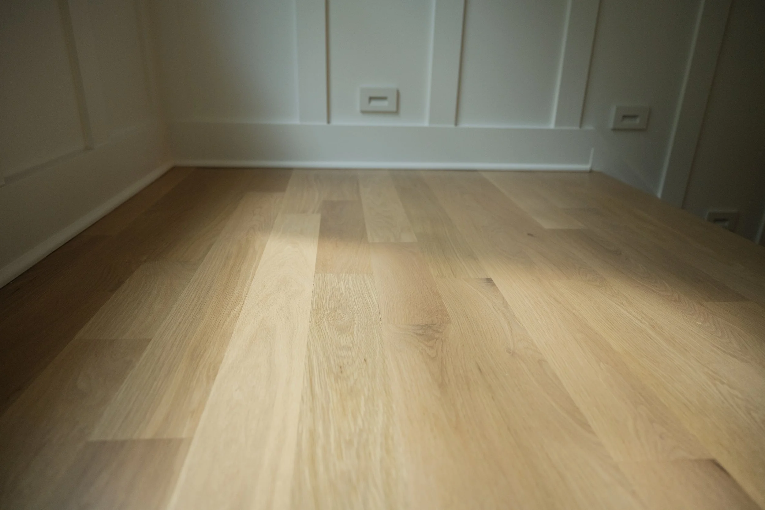 Close-up of a wooden floor against a white paneled wall with electrical outlets.