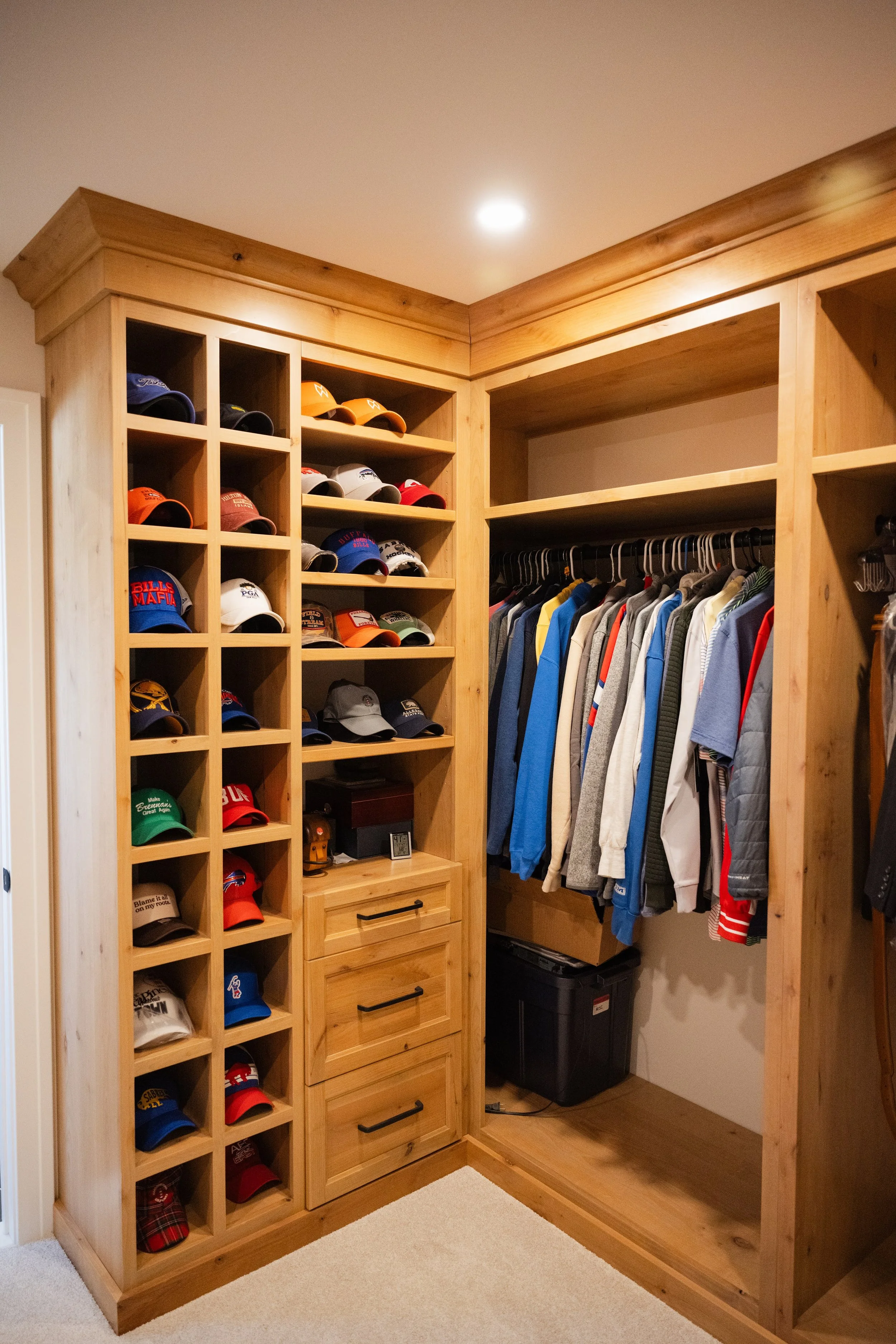 A walk-in closet with custom wooden shelving and clothing storage. The left side contains multiple cubby holes filled with various baseball caps, and below them are three drawers. On the right side, there is a hanging rod with shirts and jackets, and