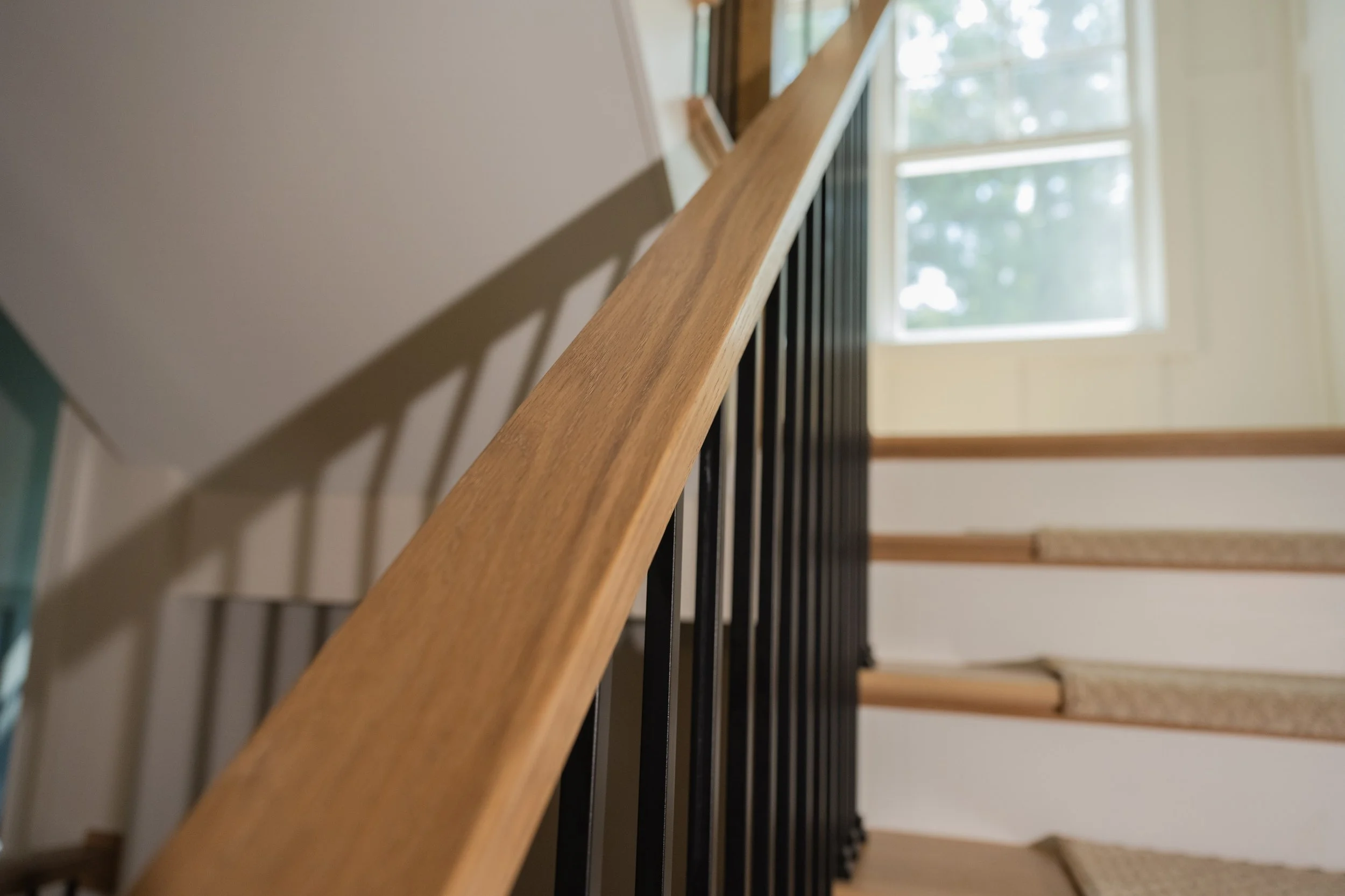Close-up of a wooden stair handrail with black balusters, viewed from below against a background of a staircase and a window letting in natural light.