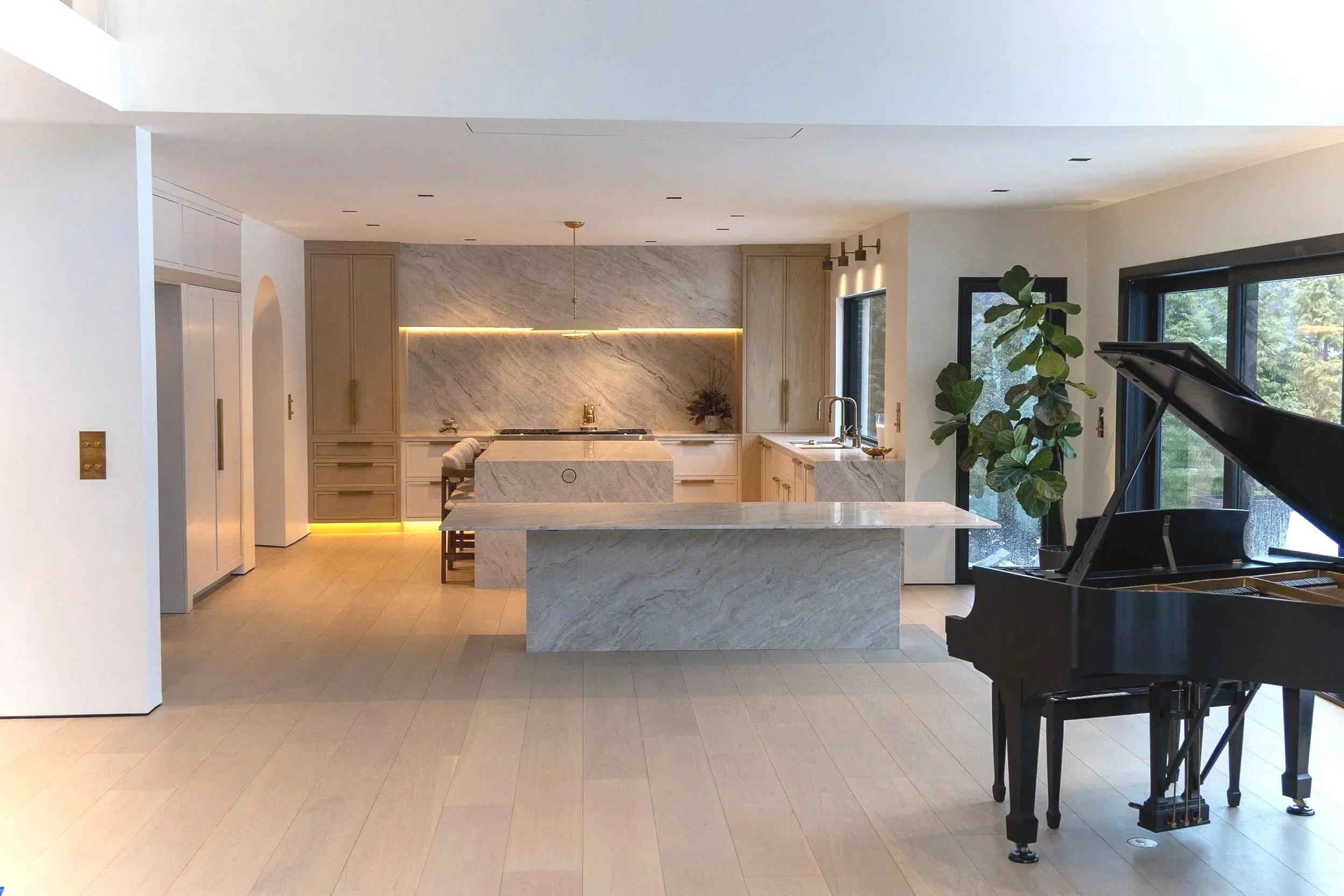 Modern kitchen with marble countertops and backsplash, light wood cabinets, large window with trees outside, a black grand piano, and a potted plant near the window.