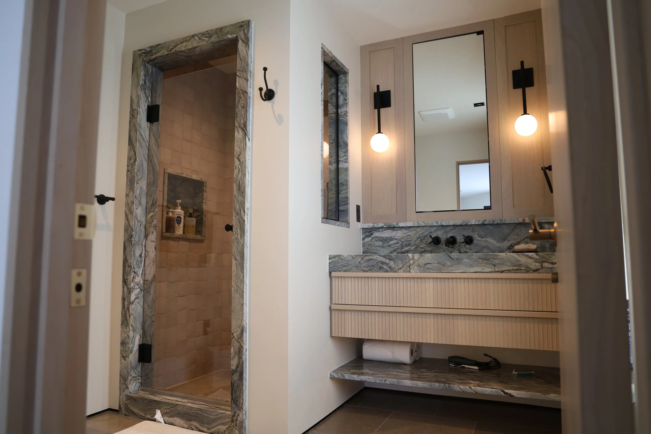 A modern bathroom with a walk-in shower featuring beige tiles and marble accents, a white vanity with a large mirror, and black fixtures on the light-colored wood cabinets. There are two spherical pendant lights hanging beside the mirror.