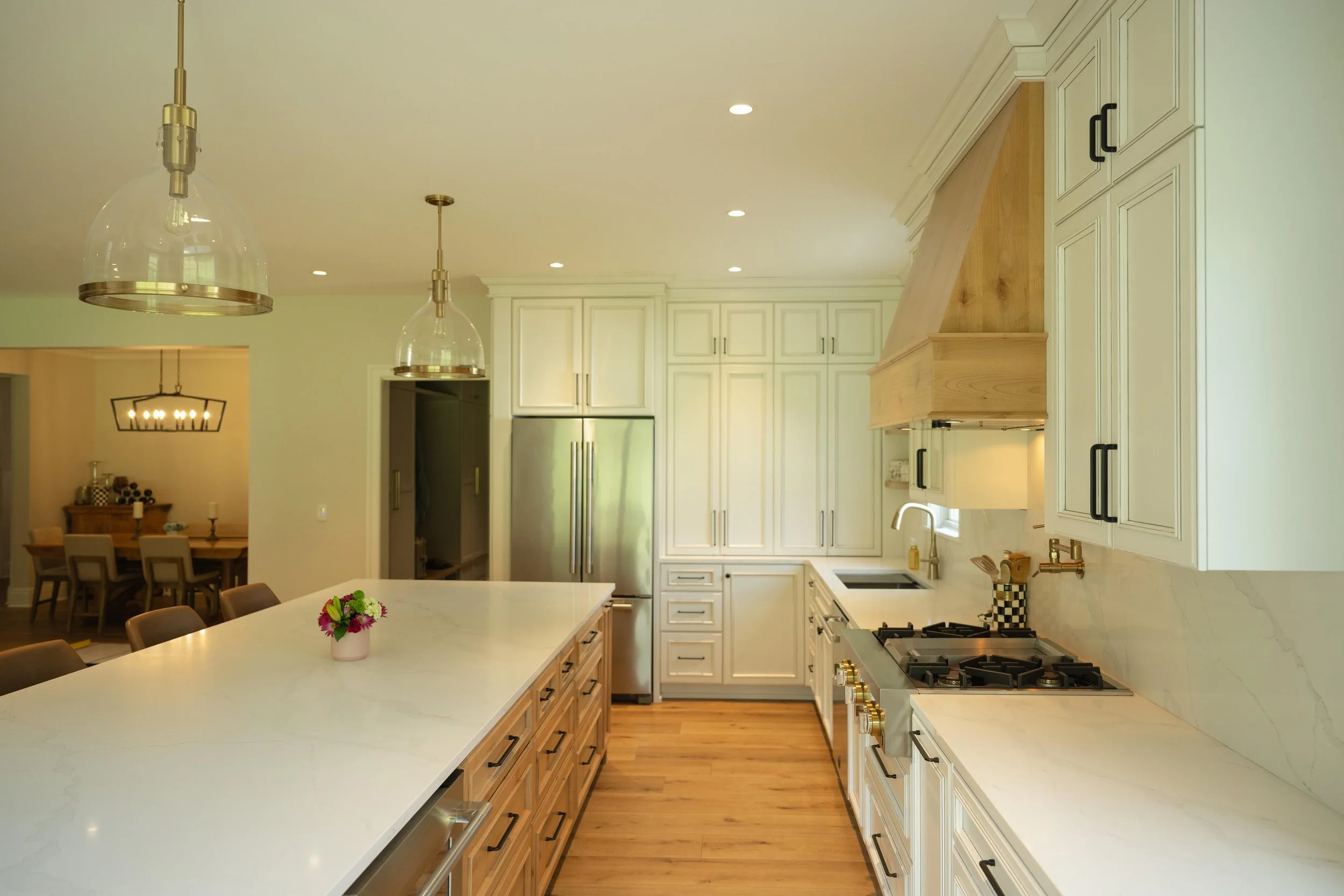 A modern kitchen with white cabinetry, a large marble island with wooden drawers, brass hardware, a stainless steel refrigerator, a gas stove, and a sink with a kitchen faucet. Recessed lighting and pendant lights hang from the ceiling. There is a di