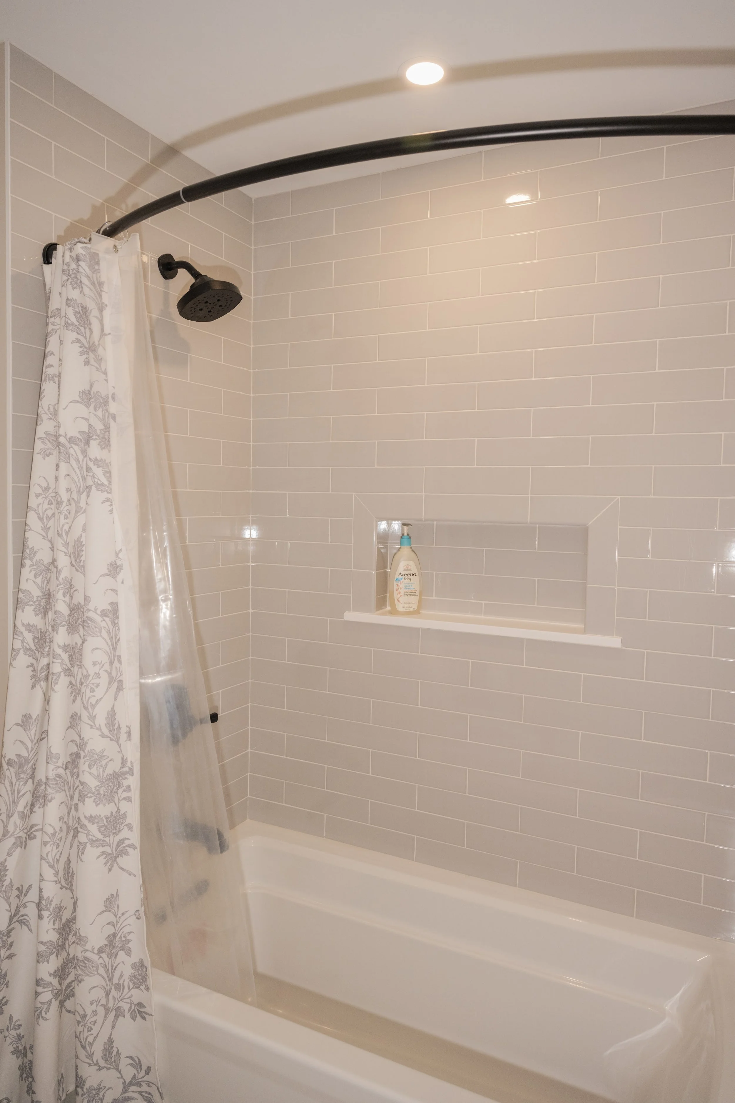 Bathroom shower area with beige tiled walls, a black showerhead, a curved shower rod with a floral-patterned shower curtain, a built-in niche with a bottle of Aveeno body wash, and a small recessed light on the ceiling.