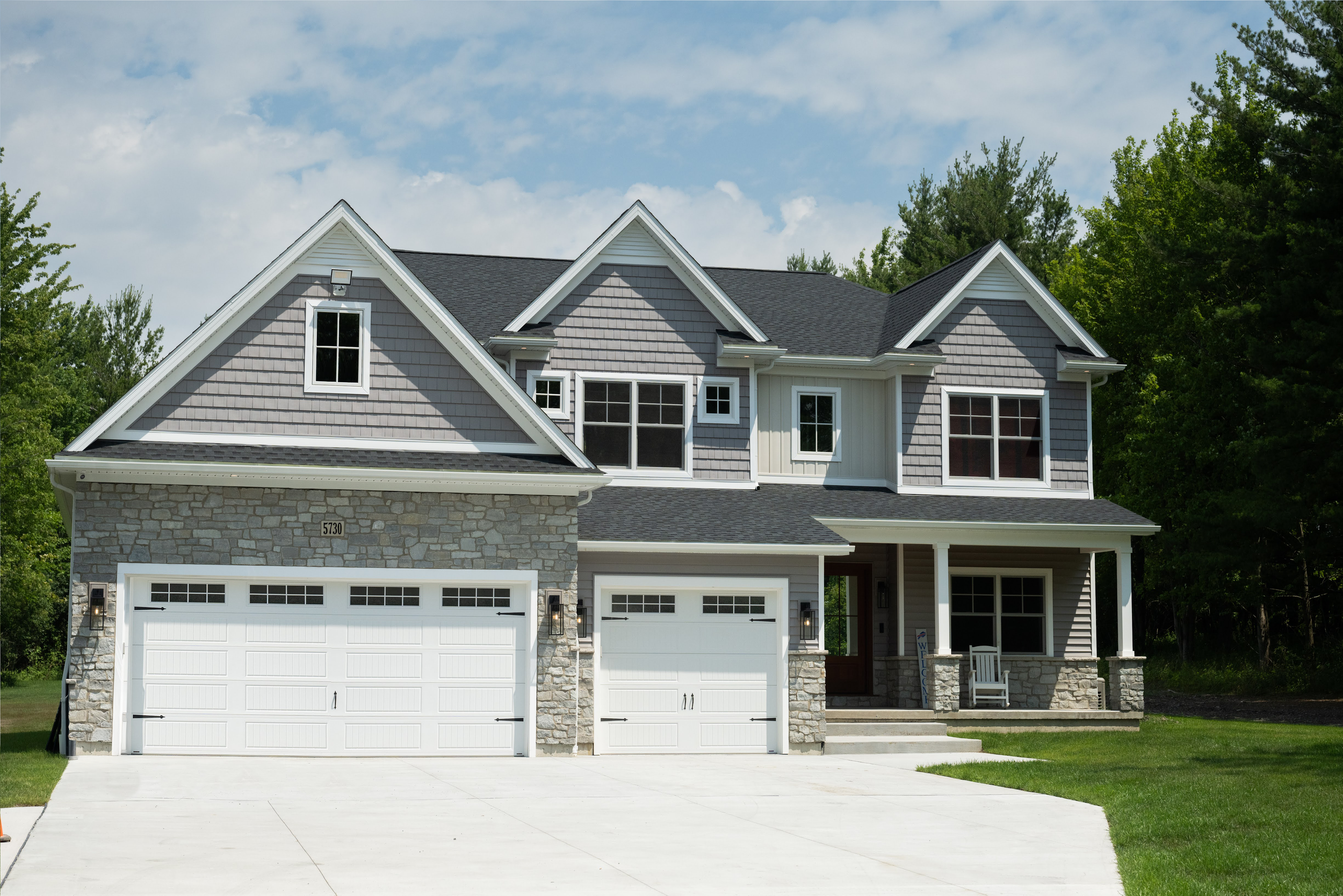Front view of a two-story house with a stone and gray siding exterior, white garage doors, and multiple gabled roofs, surrounded by green trees and a partly cloudy sky.