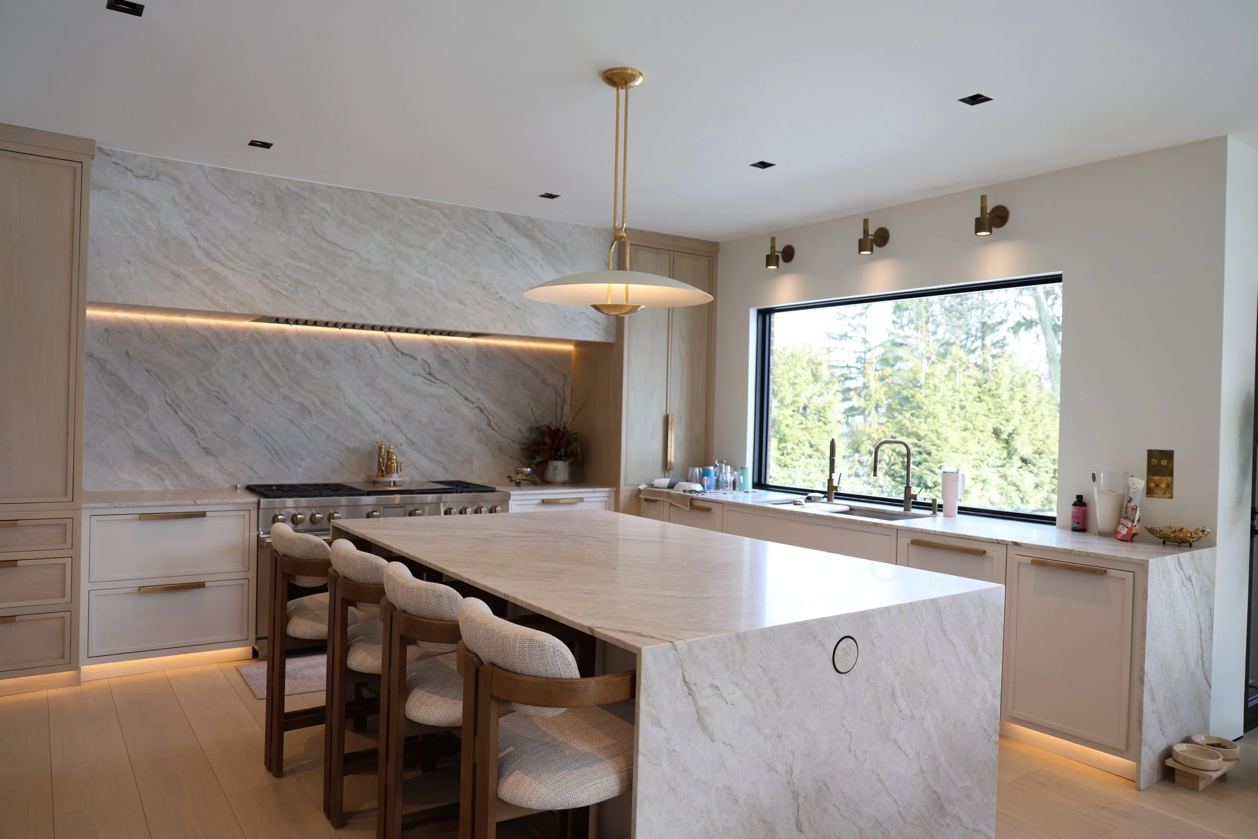Modern kitchen with light wood cabinetry, a marble island, a large window, and minimalist pendant lighting.