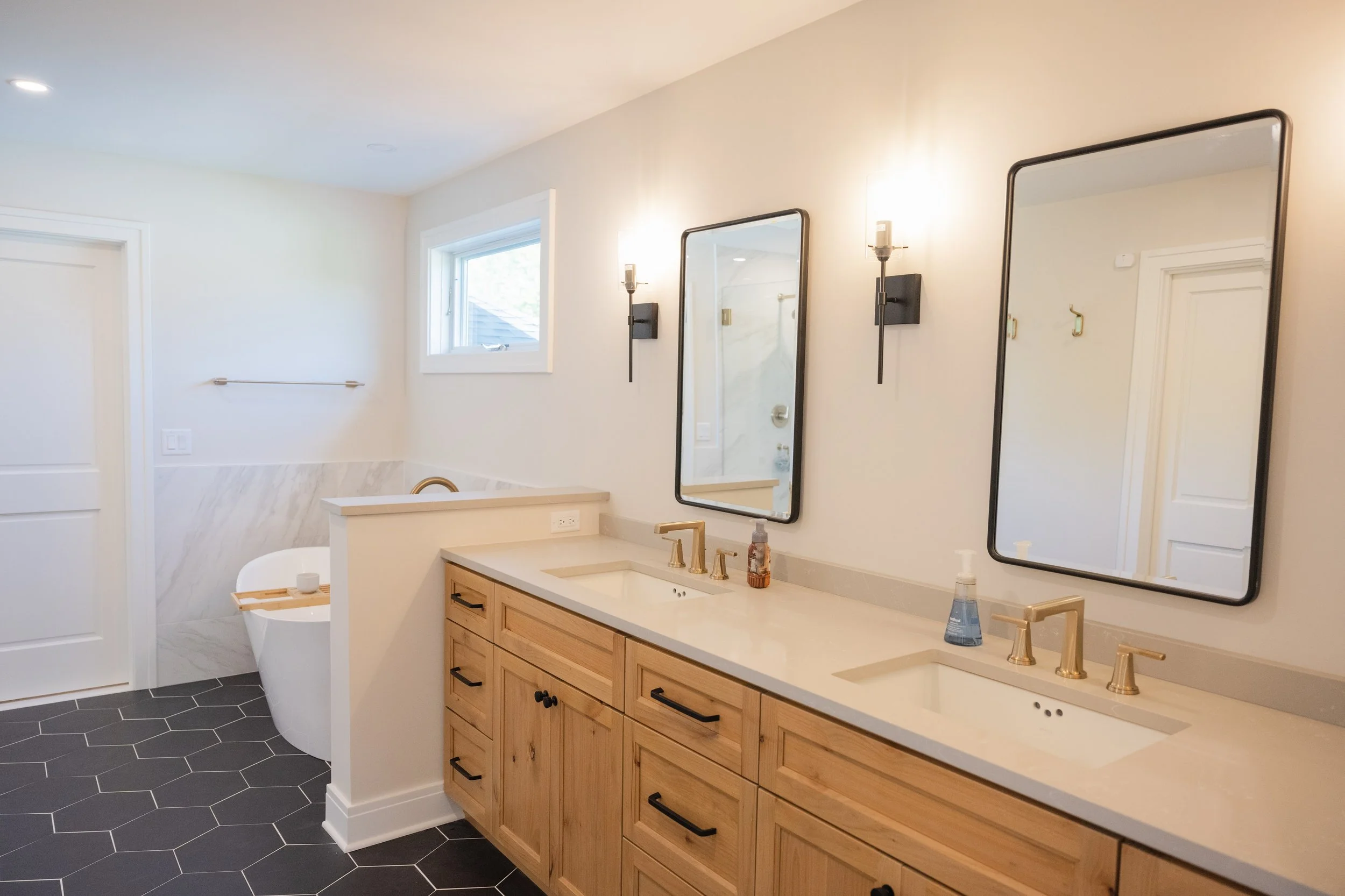 A modern bathroom with a double vanity featuring wooden cabinets, gold fixtures, and two rectangular mirrors. Light walls and natural light from a window brighten the space. A partial view of a shower with marble tiles and a freestanding bathtub with