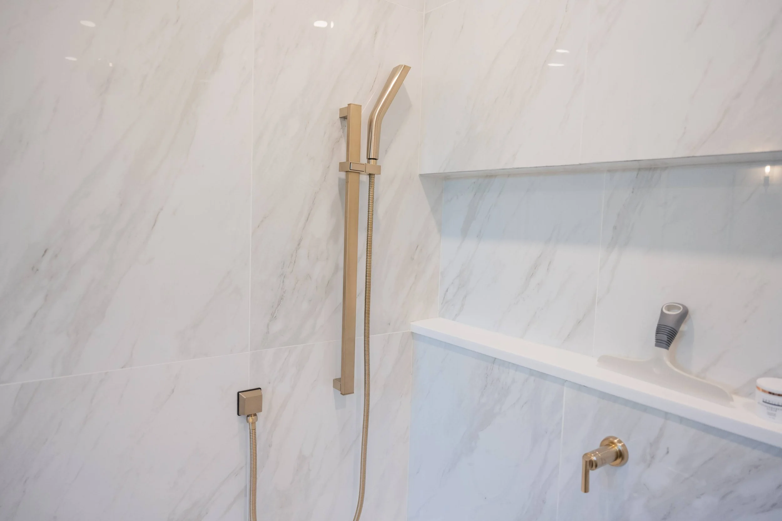 A shower area with a beige handheld showerhead mounted on a vertical bar on a marble wall, with a built-in shelf holding a bottle of shampoo or body wash, and a small gray shower head on the wall.