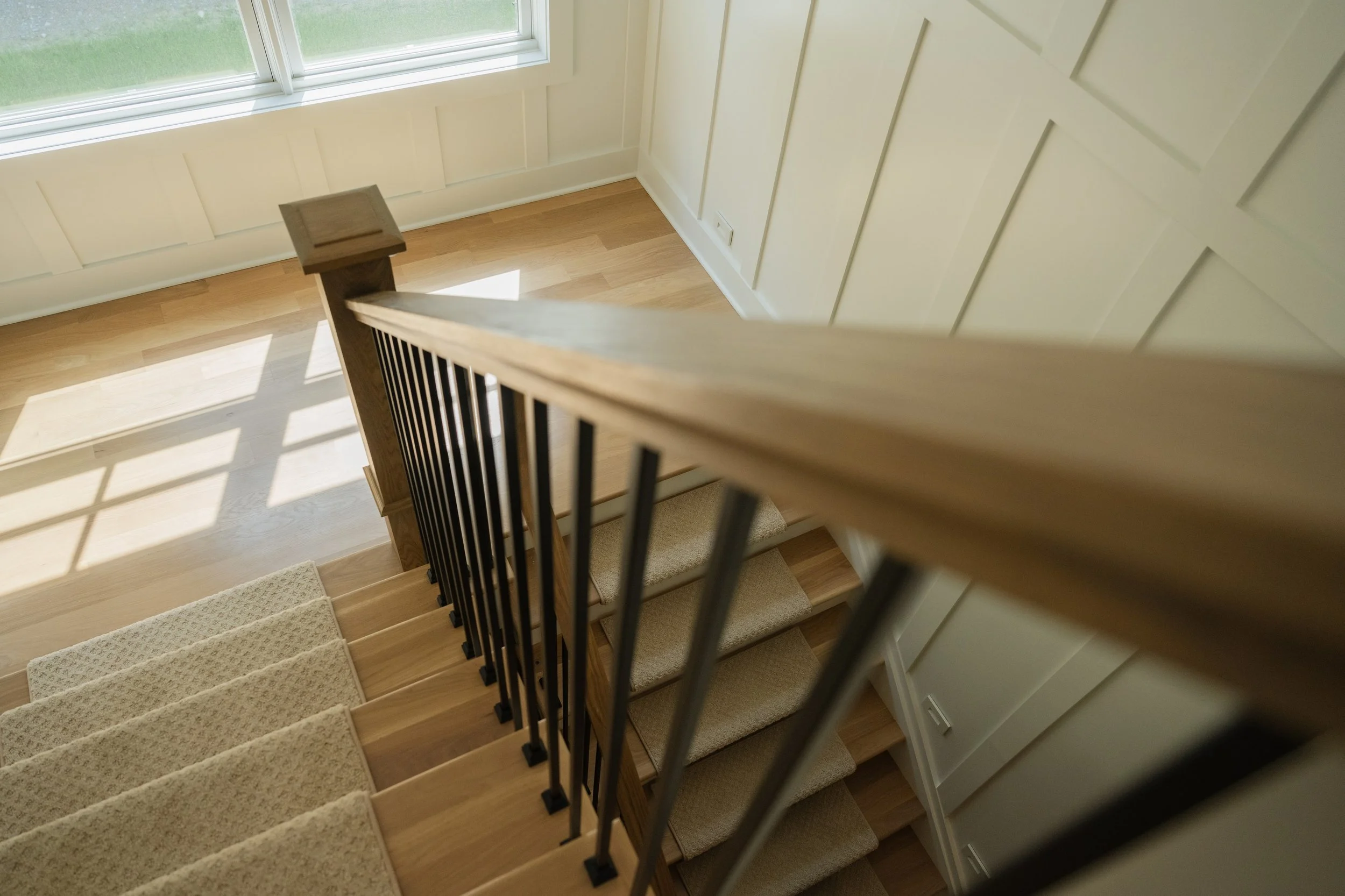 View of a staircase from above near a window, with wooden steps, black metal railing, and beige carpet runner on the stairs.