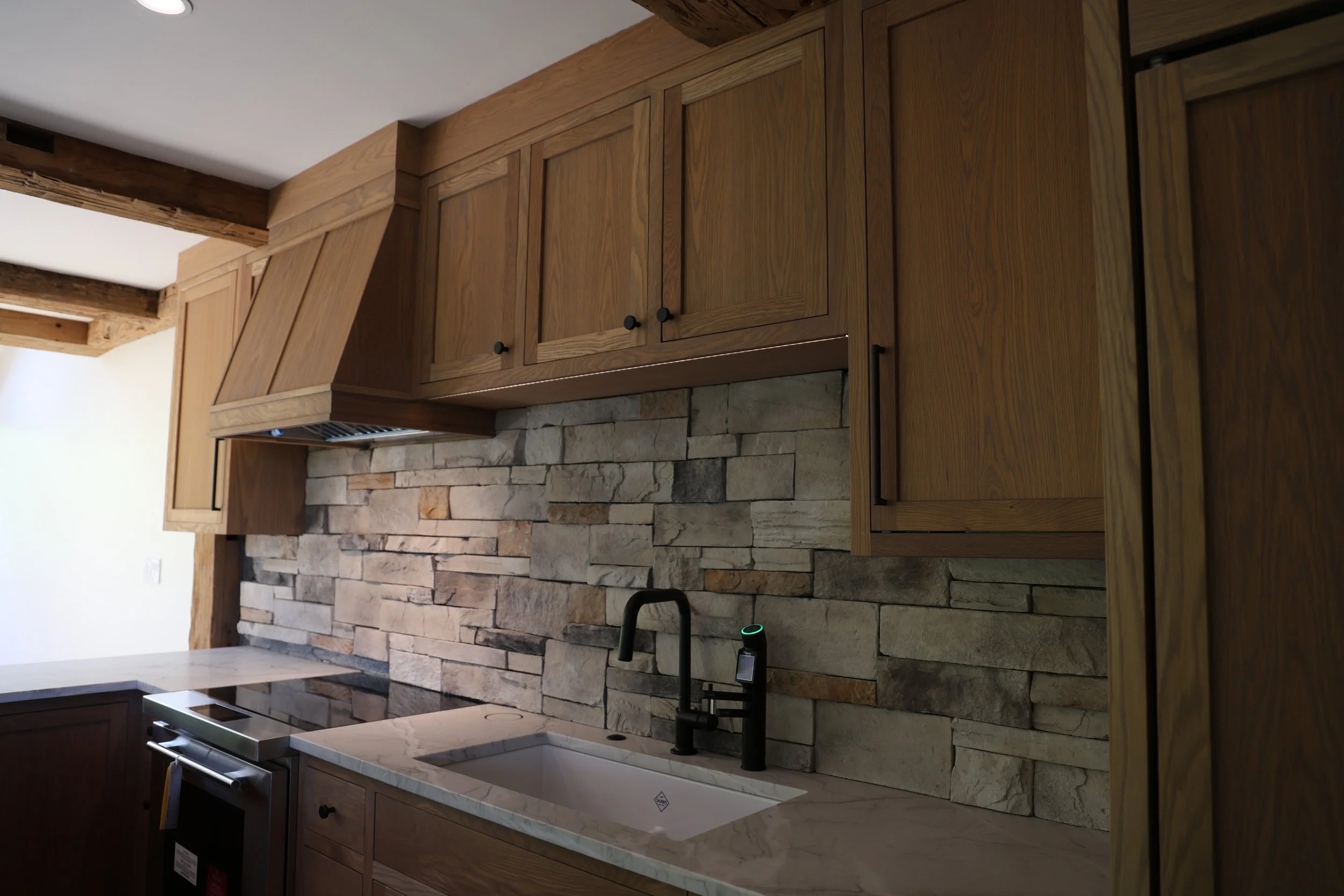 Kitchen with wooden cabinets, stone backsplash, and a white farmhouse sink with a matte black faucet in Ellicottville, NY.