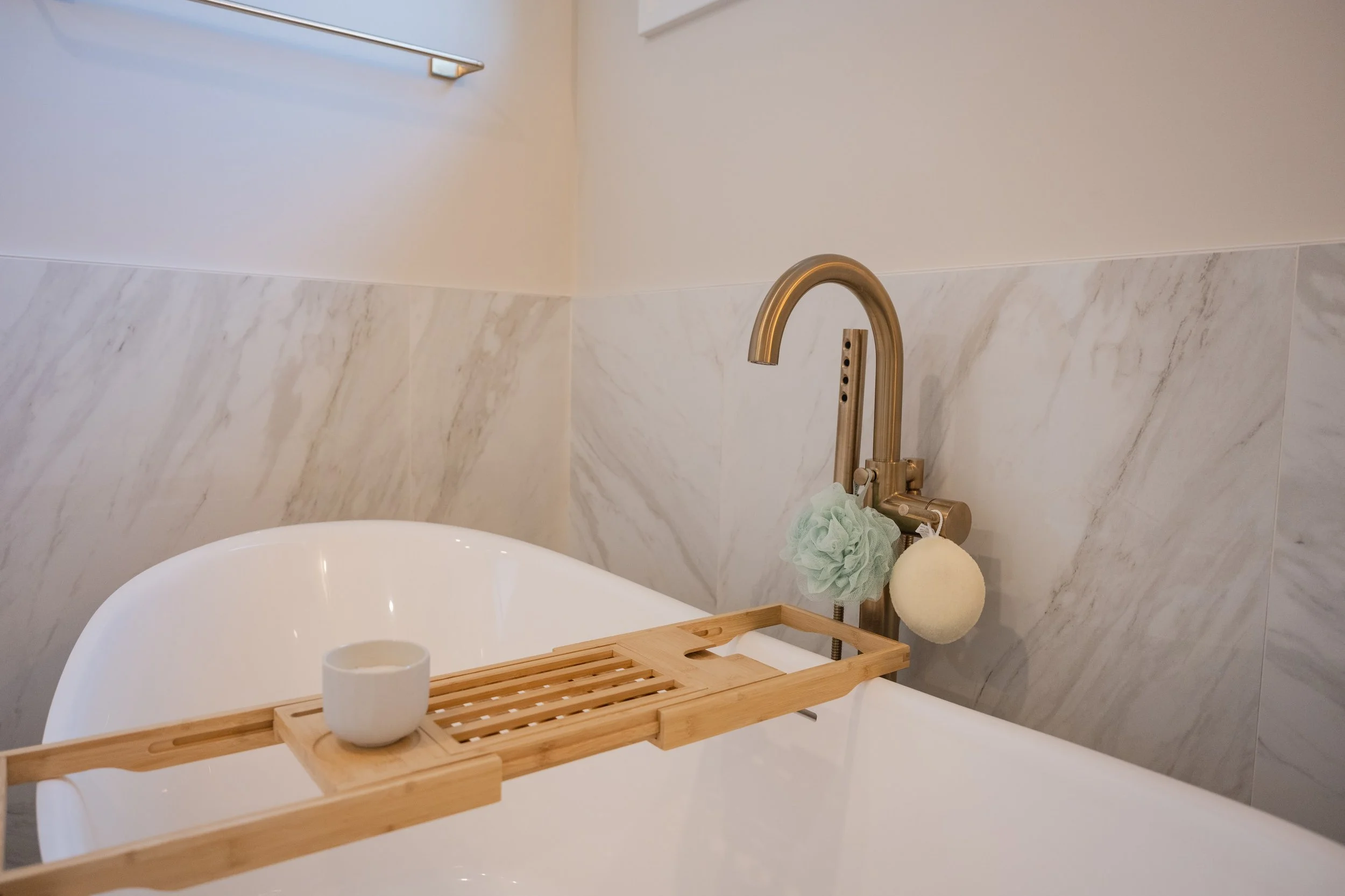 A modern bathtub with a wooden bath tray holding a white cup, with a gold faucet and a bath sponge hanging on the wall.