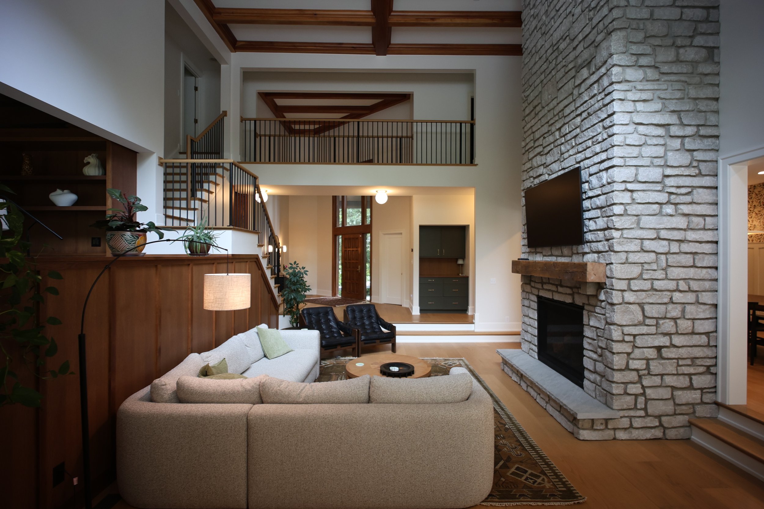 Interior view of a living room with a large stone fireplace, beige sectional sofa, black chairs, wooden staircase, and high ceiling with wooden beams.