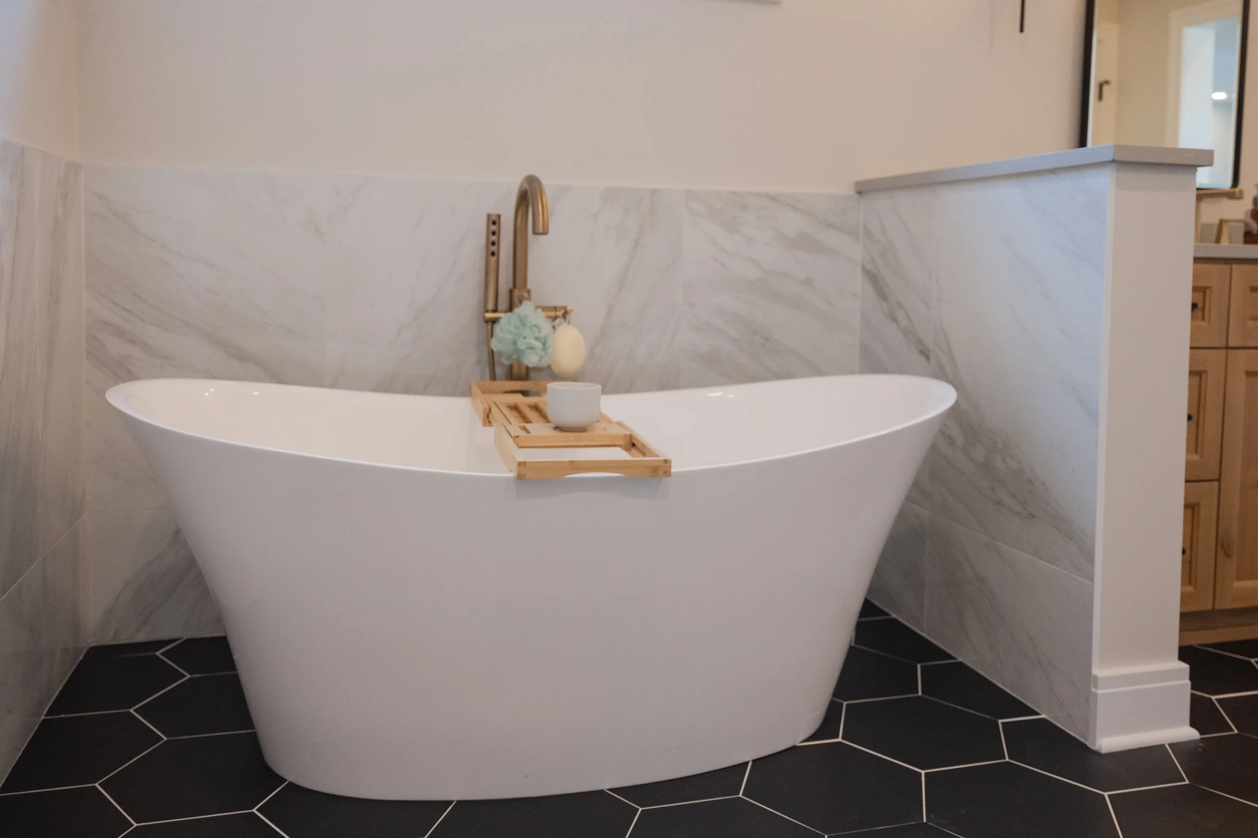 A modern bathroom with a white freestanding bathtub placed on black hexagonal floor tiles. The background features a wall with marble panels and a brass faucet with a spa sponge and bath brush hanging from it. Wooden furniture and a mirror are visibl