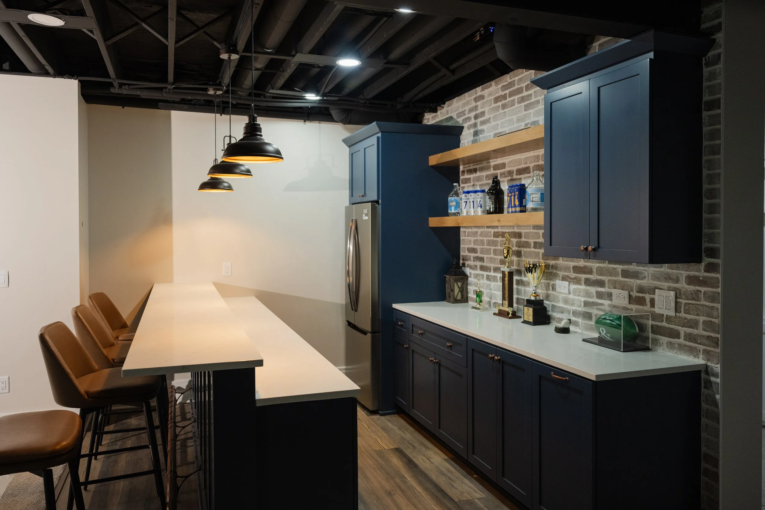 Interior view of a modern kitchen with black exposed ceiling beams, dark blue cabinets, and a brick wall. There is a long table with brown chairs, pendant lights hanging from the ceiling, and various trophies displayed on the countertop.