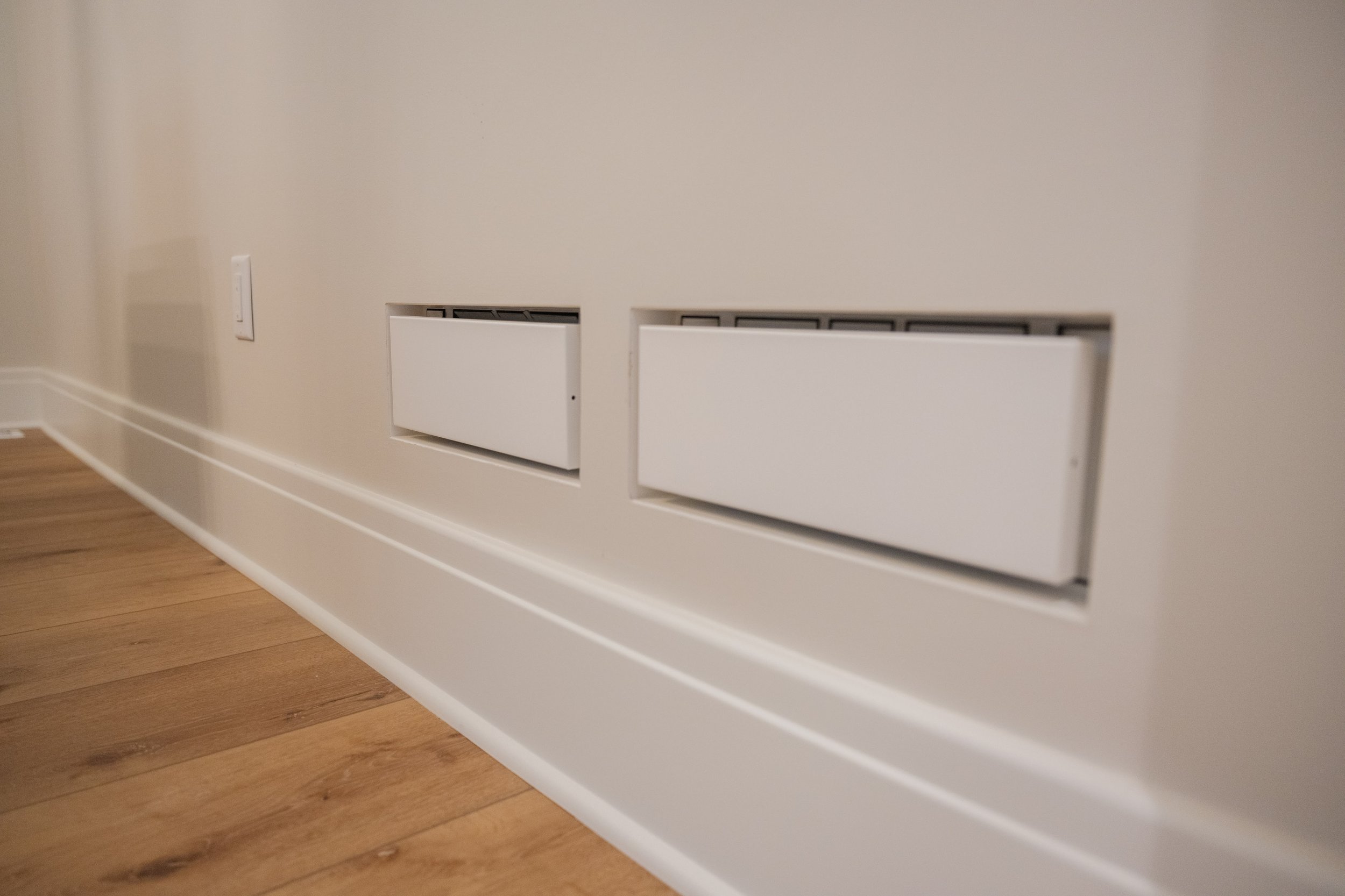 Close-up of a white air vent cover on a beige wall, with a power outlet visible to the left and a wooden floor below.