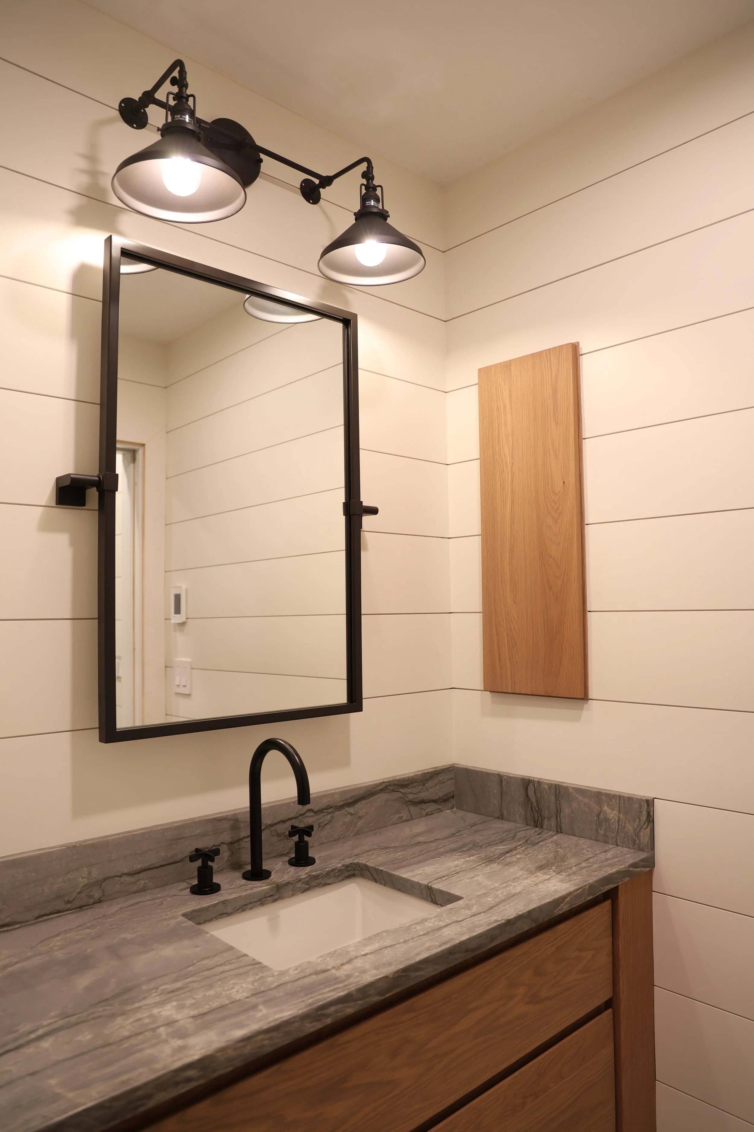 A bathroom ledge with a gray marble countertop, a black faucet, a rectangular mirror with a black frame, a wooden wall panel, and overhead industrial-style light fixtures.