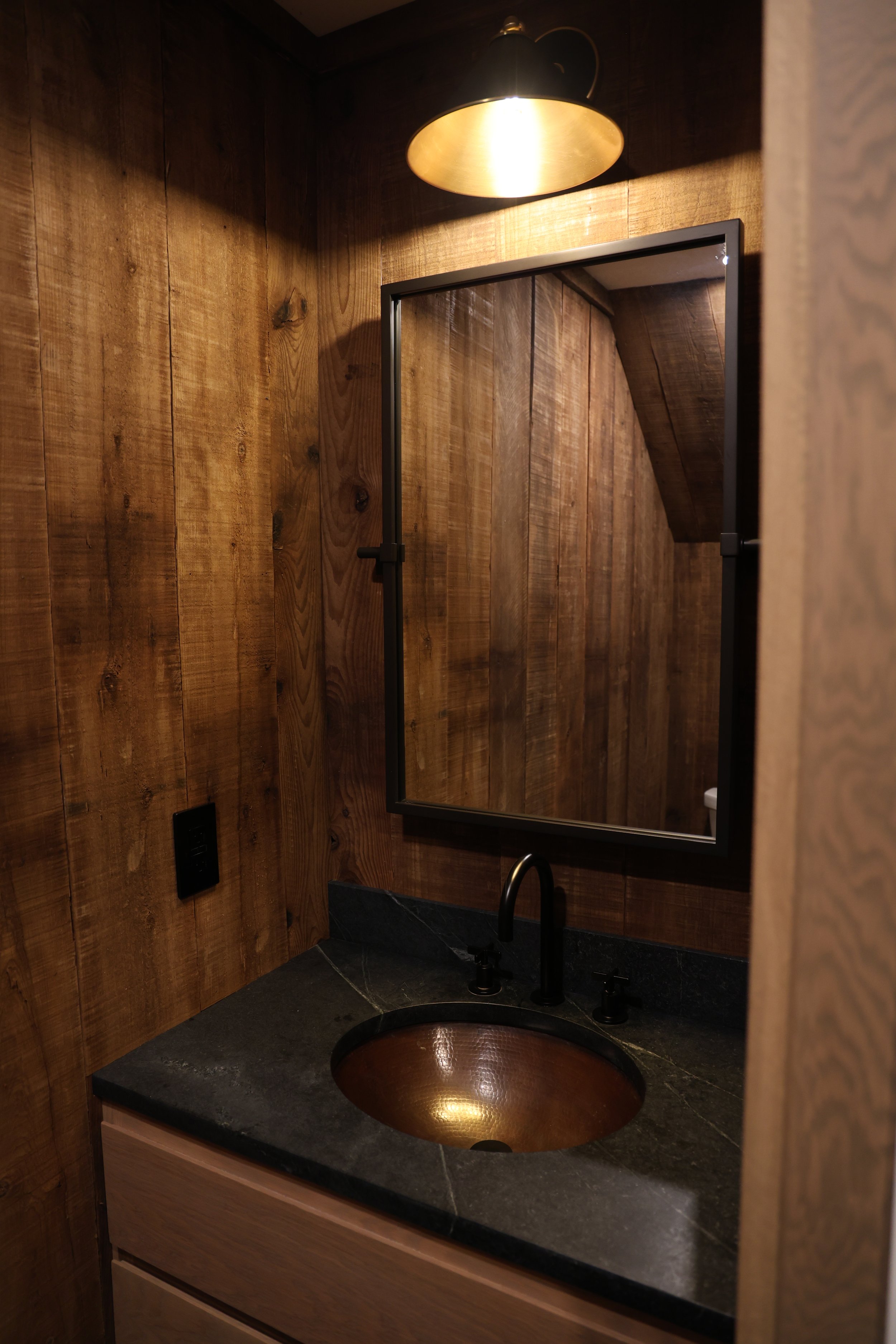 A bathroom with wooden paneled walls, a black countertop with a copper-colored sink, a mirror, and a black faucet. A black and gold ceiling light is reflected in the mirror.