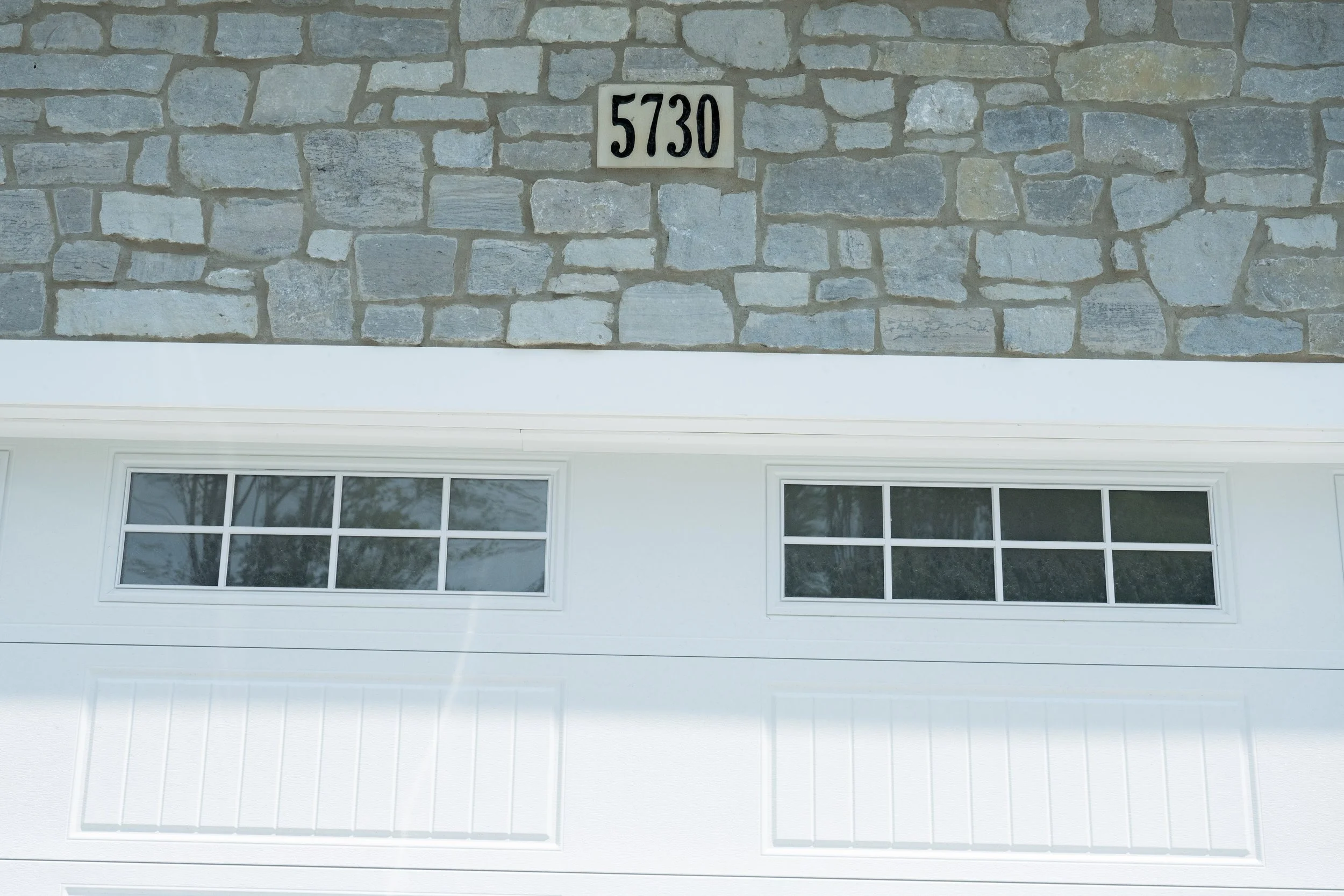 Exterior of a house showing a stone facade with a house number sign reading '5730' above two white-framed windows.