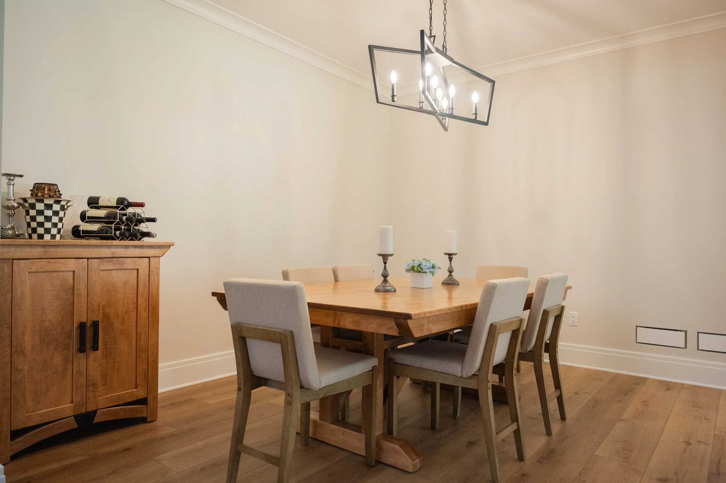 Interior of a dining room with a wooden table surrounded by six beige upholstered chairs, a wooden sideboard on the left with bottles of wine and decorative items, and a hanging modern chandelier with exposed bulbs. There are two candles and a small 