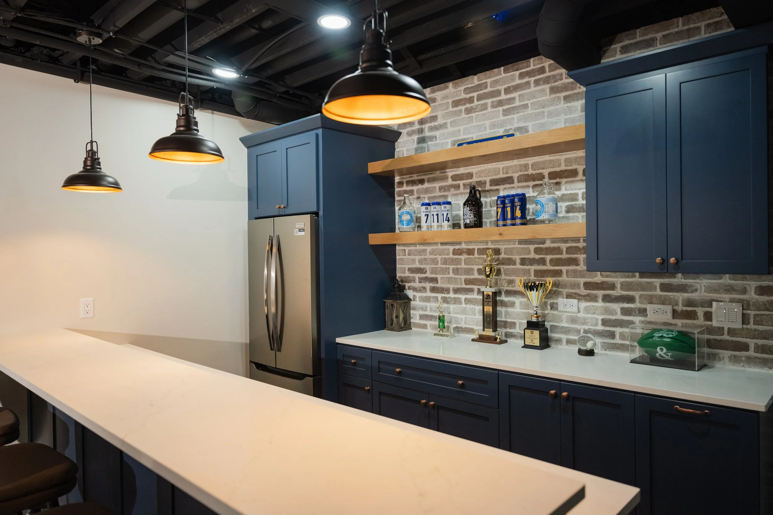 Modern kitchen with navy blue cabinets, brick backsplash, open wooden shelves, stainless steel refrigerator, black pendant lights, and display of trophies and decorative items on the counter.