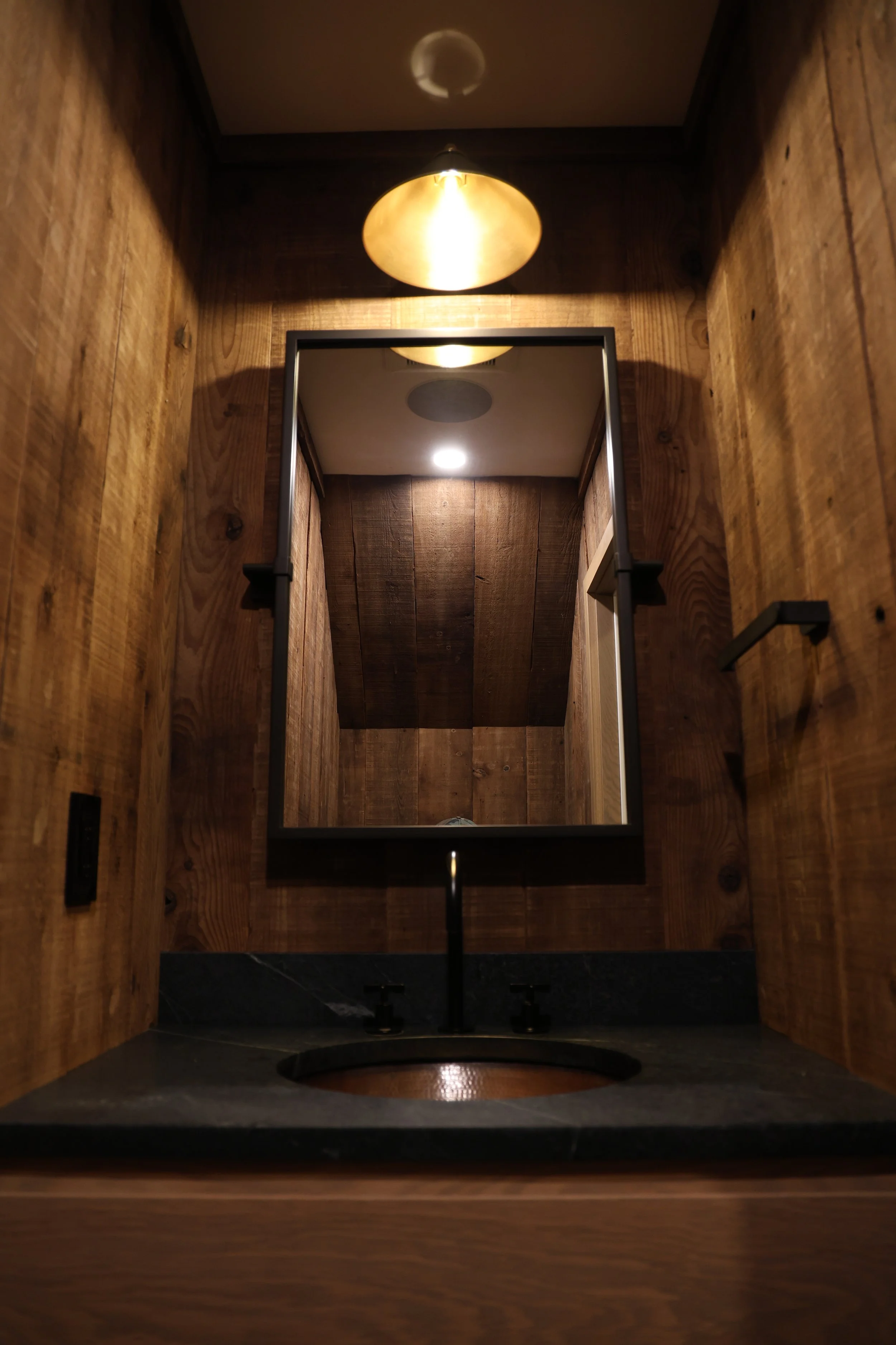 A mirror mounted on wood-paneled wall reflecting ceiling lights and wooden ceiling beams, with a bathroom sink and faucet visible below