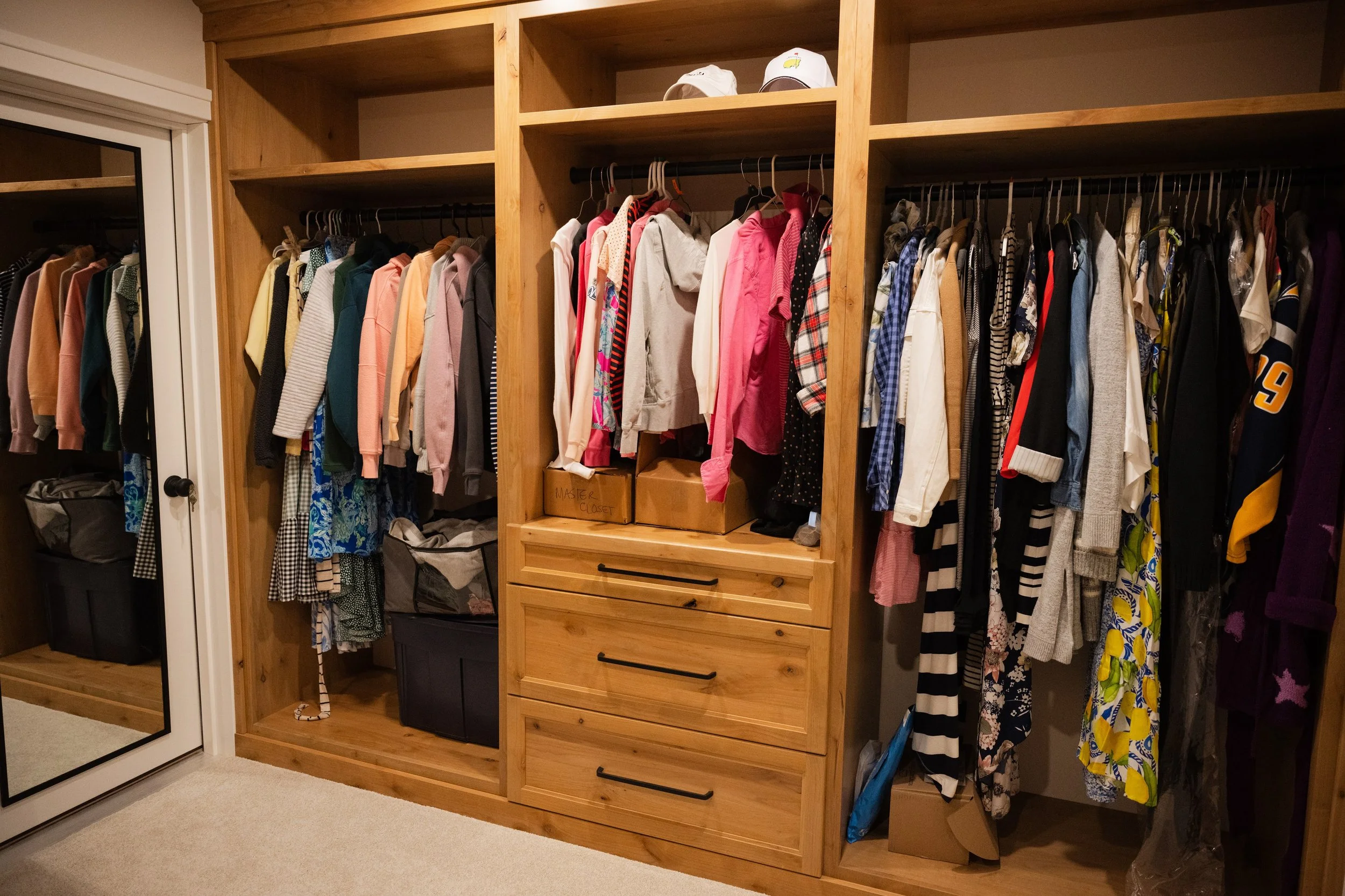A closet with wooden shelving and hanging rods filled with various clothes, shoes, and accessories, with a mirror on the left side.
