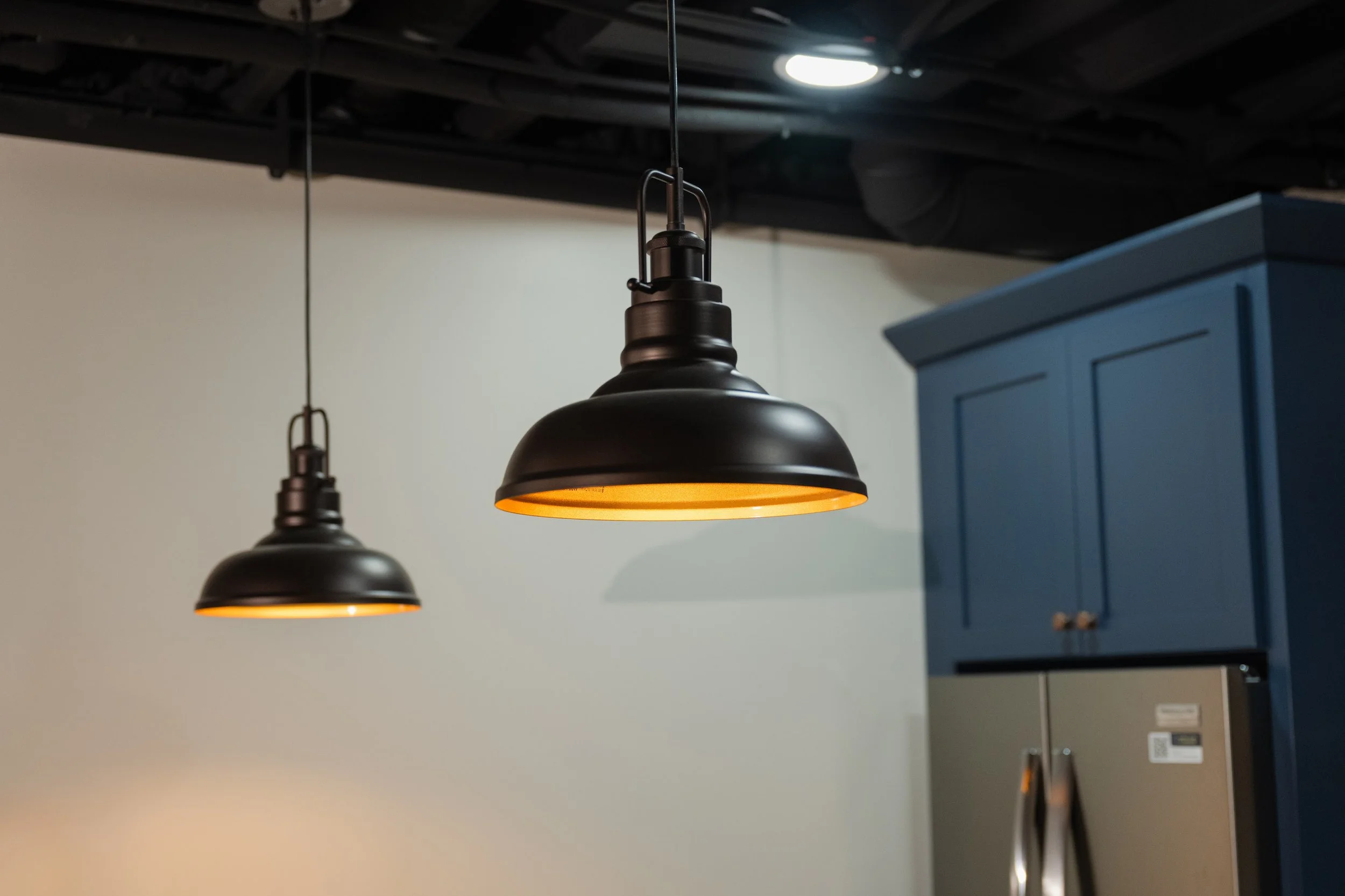Black hanging pendant light with warm yellow light in a modern kitchen with blue cabinets and a white wall.