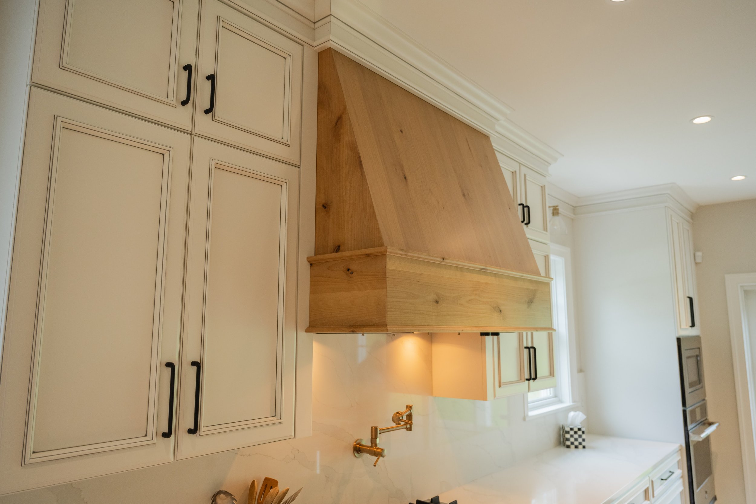 A kitchen with white cabinets, black handles, a wooden vent hood, and a white marble countertop with a gold faucet.