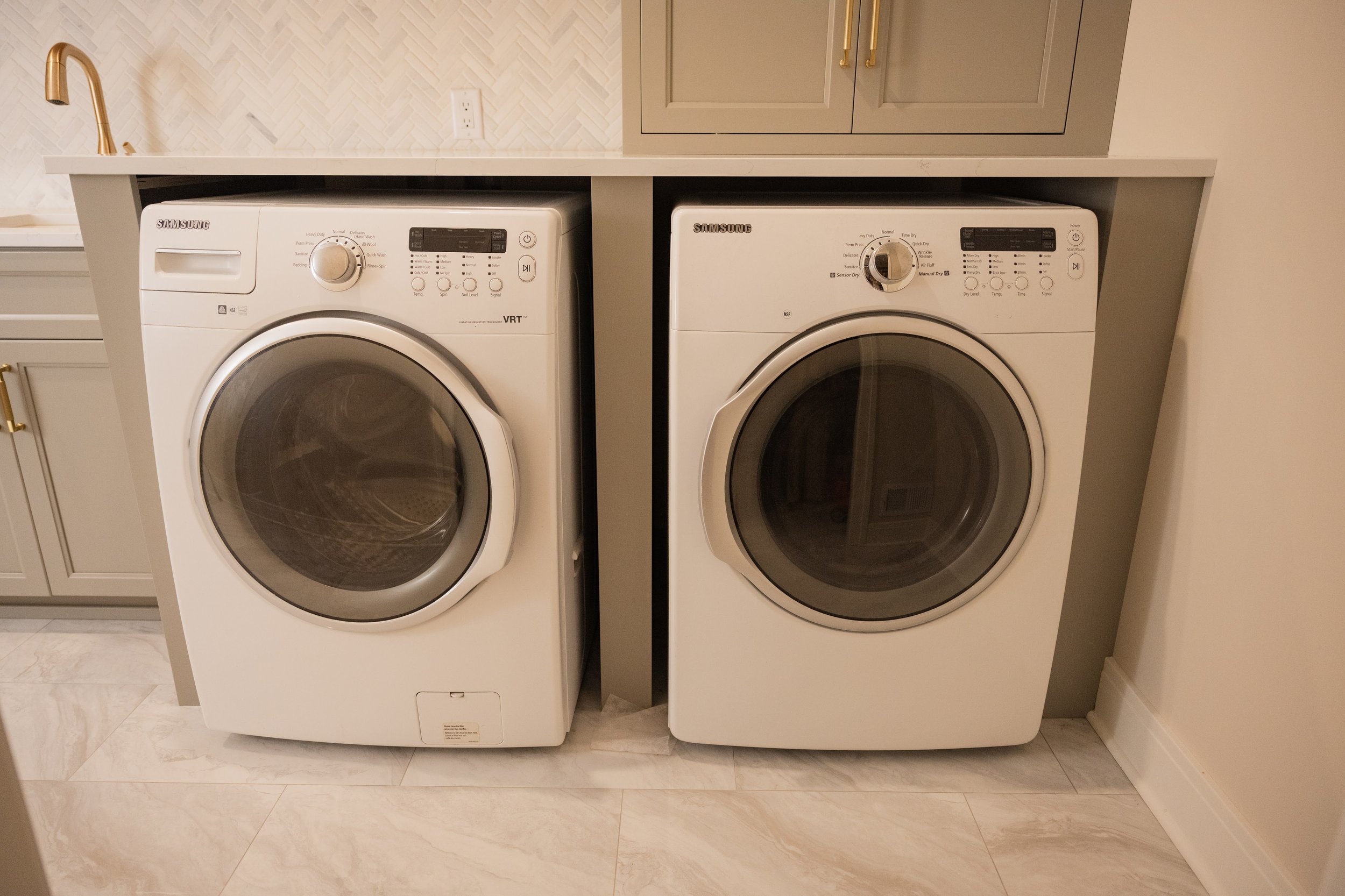 A side-by-side view of a white Samsung washing machine and dryer in a laundry room with beige cabinets and tile flooring.