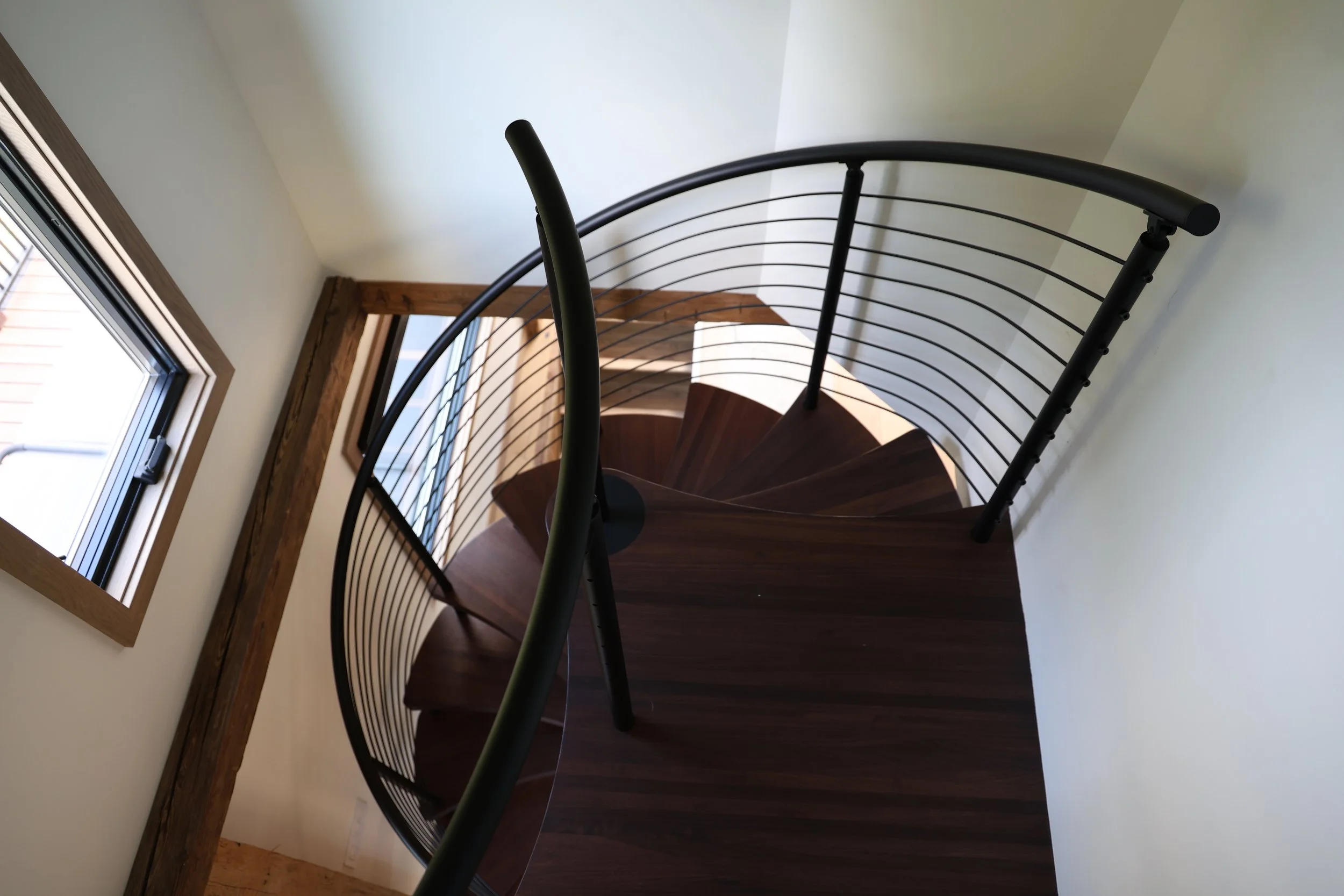 A spiral staircase with wooden steps and black metal railings seen from above, near a window with a wooden frame and a white wall.
