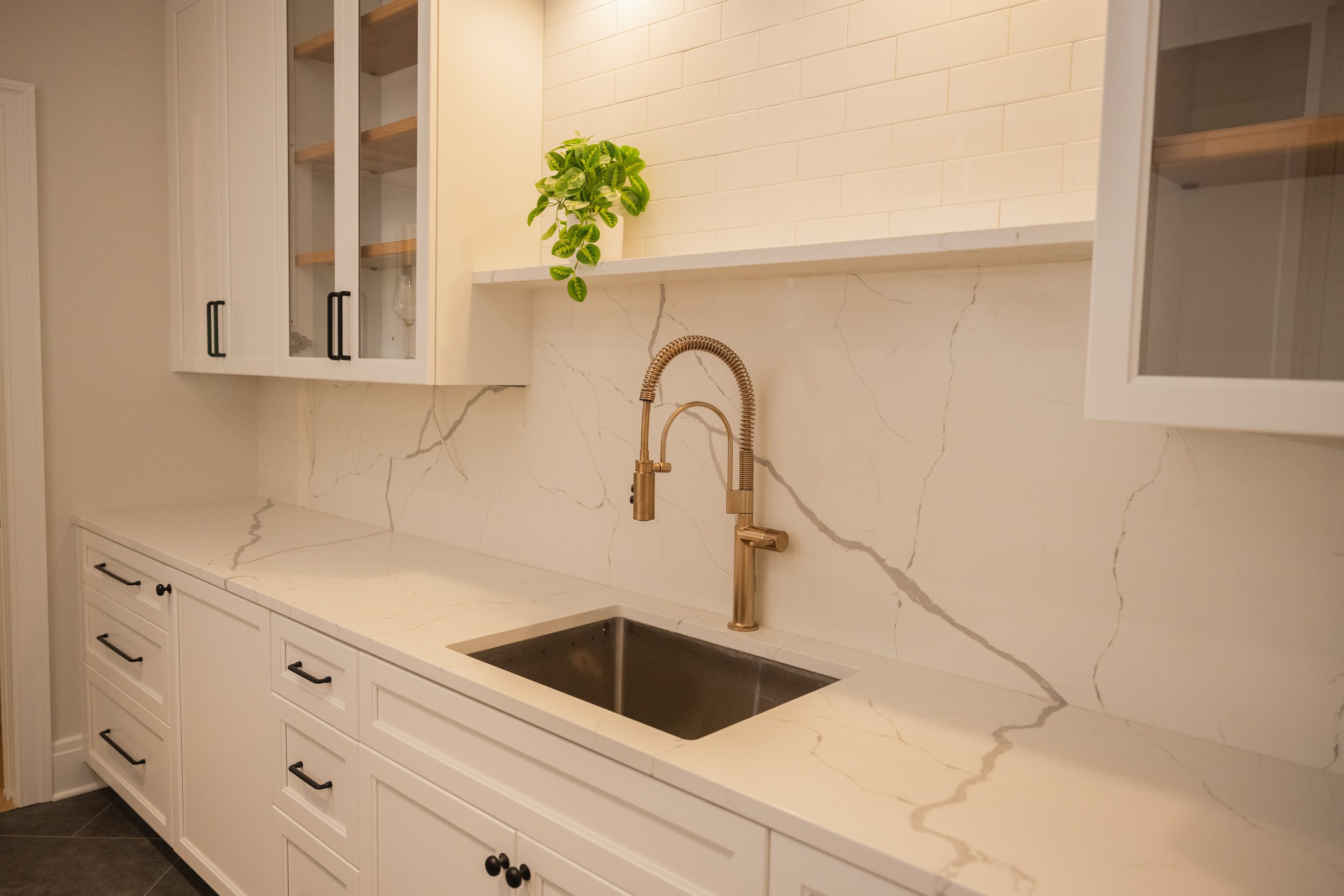 A modern kitchen with white cabinetry, a marble countertop, a matte black faucet, and a green potted plant on a shelf.