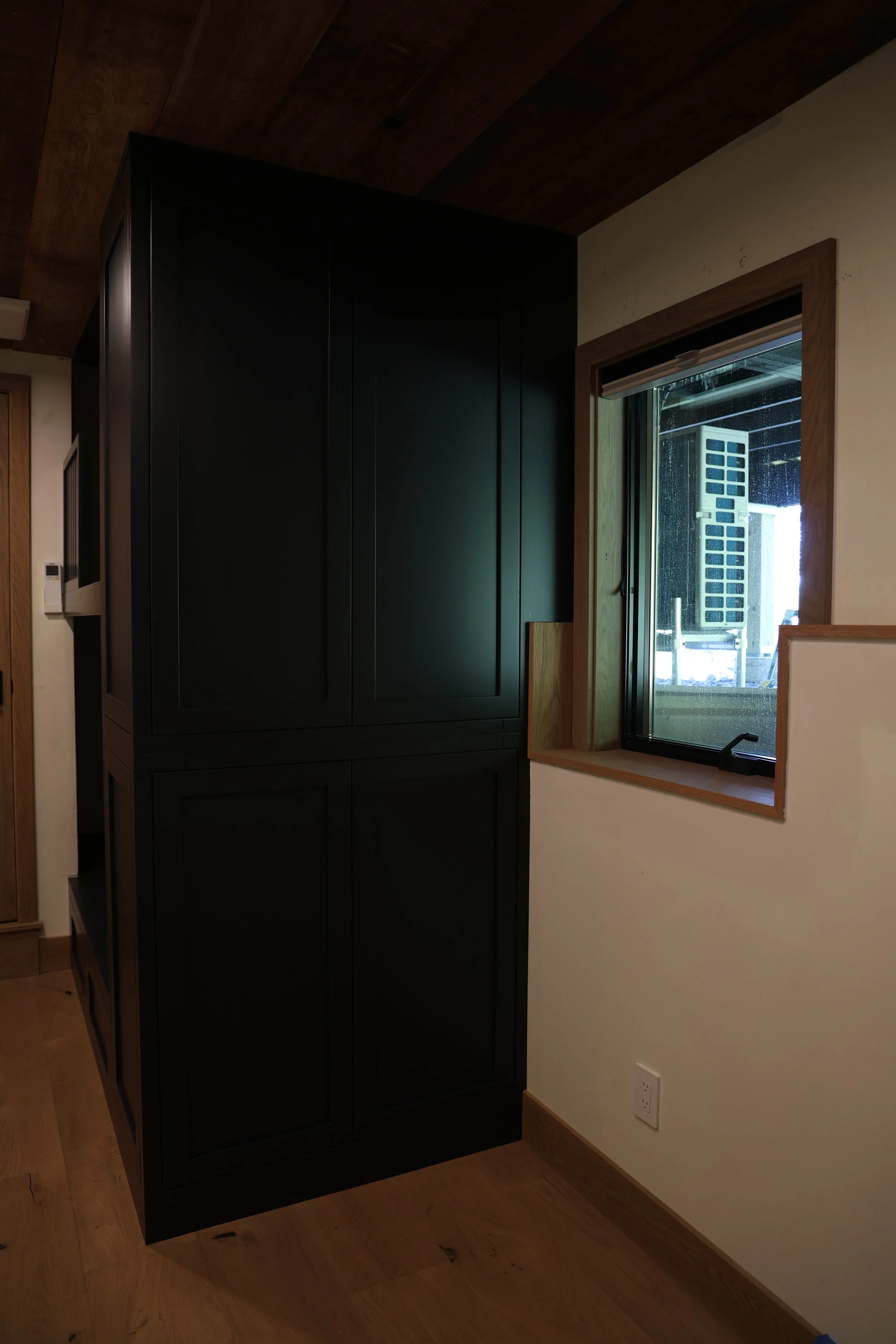 Interior view of a room featuring a large black cabinet next to a window with a wooden frame, showing an outdoor air conditioning unit. The room has a wooden ceiling and flooring, with a white wall and electrical outlet visible.
