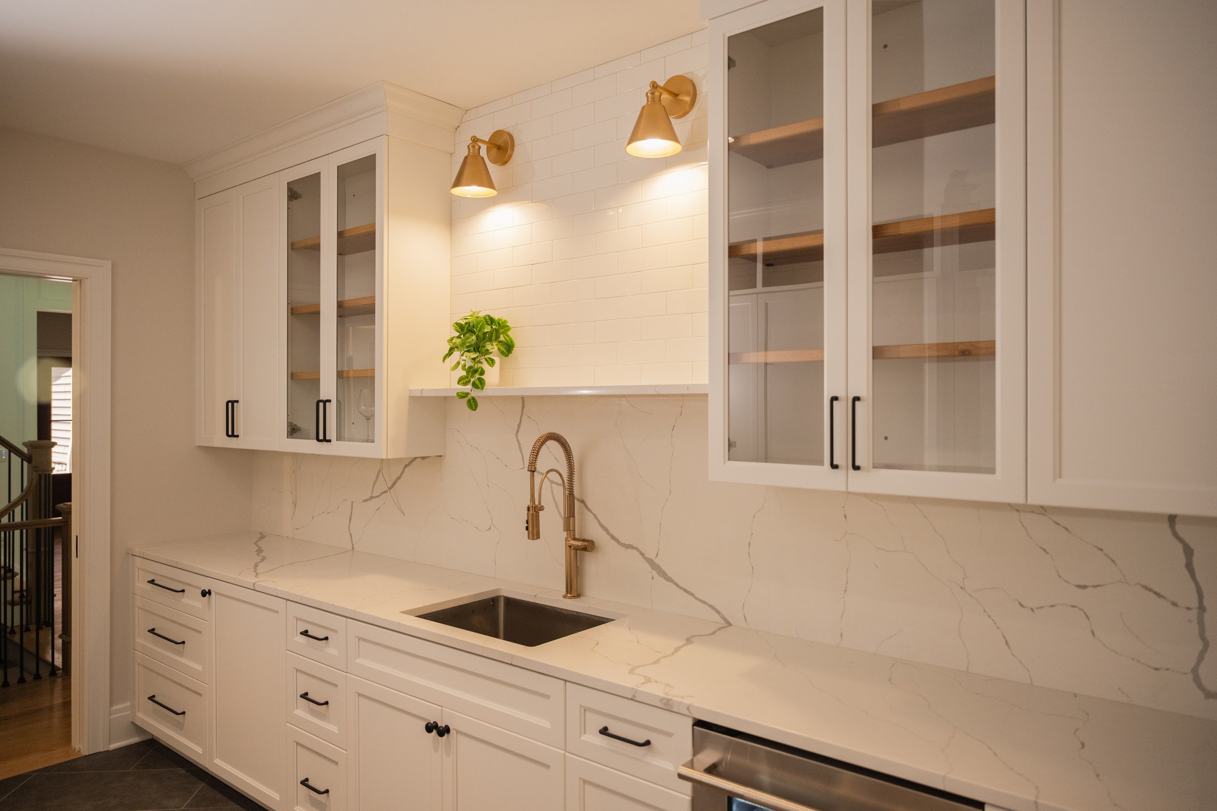 Kitchen with white cabinetry, marble countertop, and backsplash, gold wall lights, a plant, and a sink with a bronze faucet.