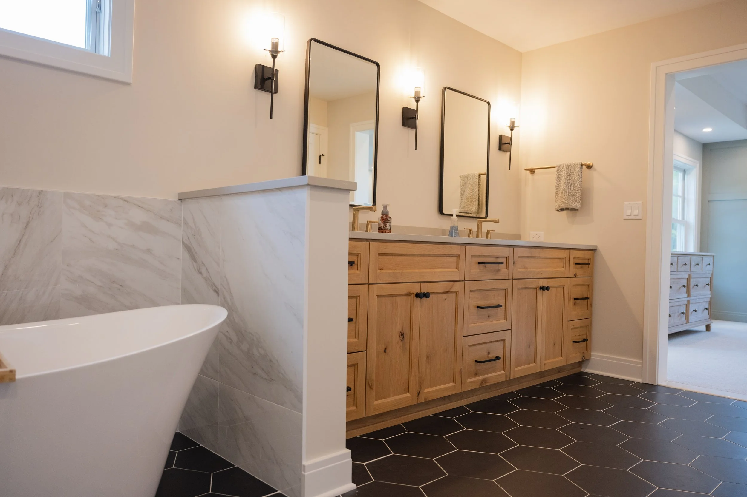 Bathroom with wooden vanity, large wall mirrors, black wall-mount light fixtures, and black hexagonal floor tiles.