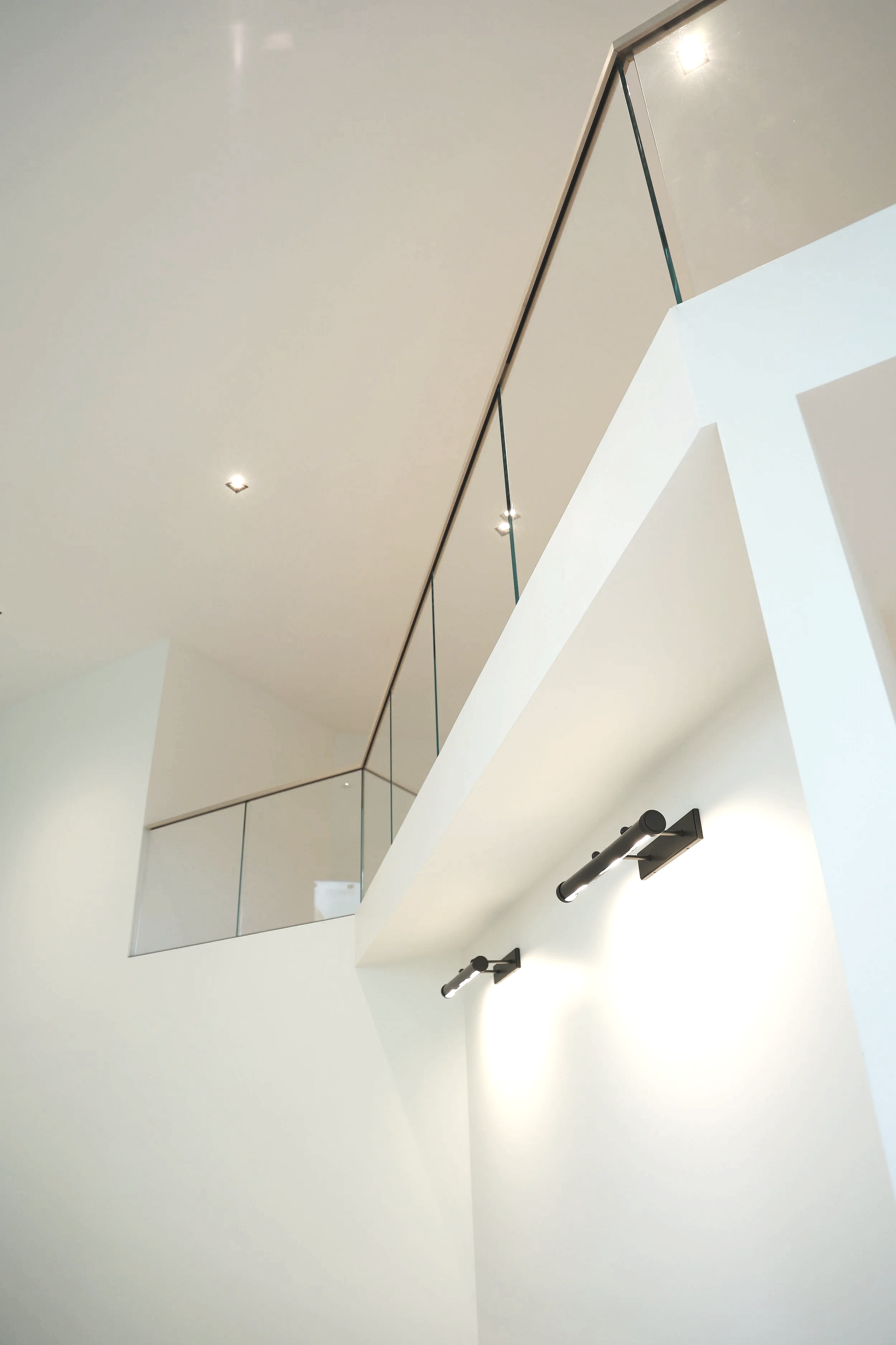 Interior view of a modern, minimalist staircase with white walls, black wall-mounted lights, and a glass and metal railing.