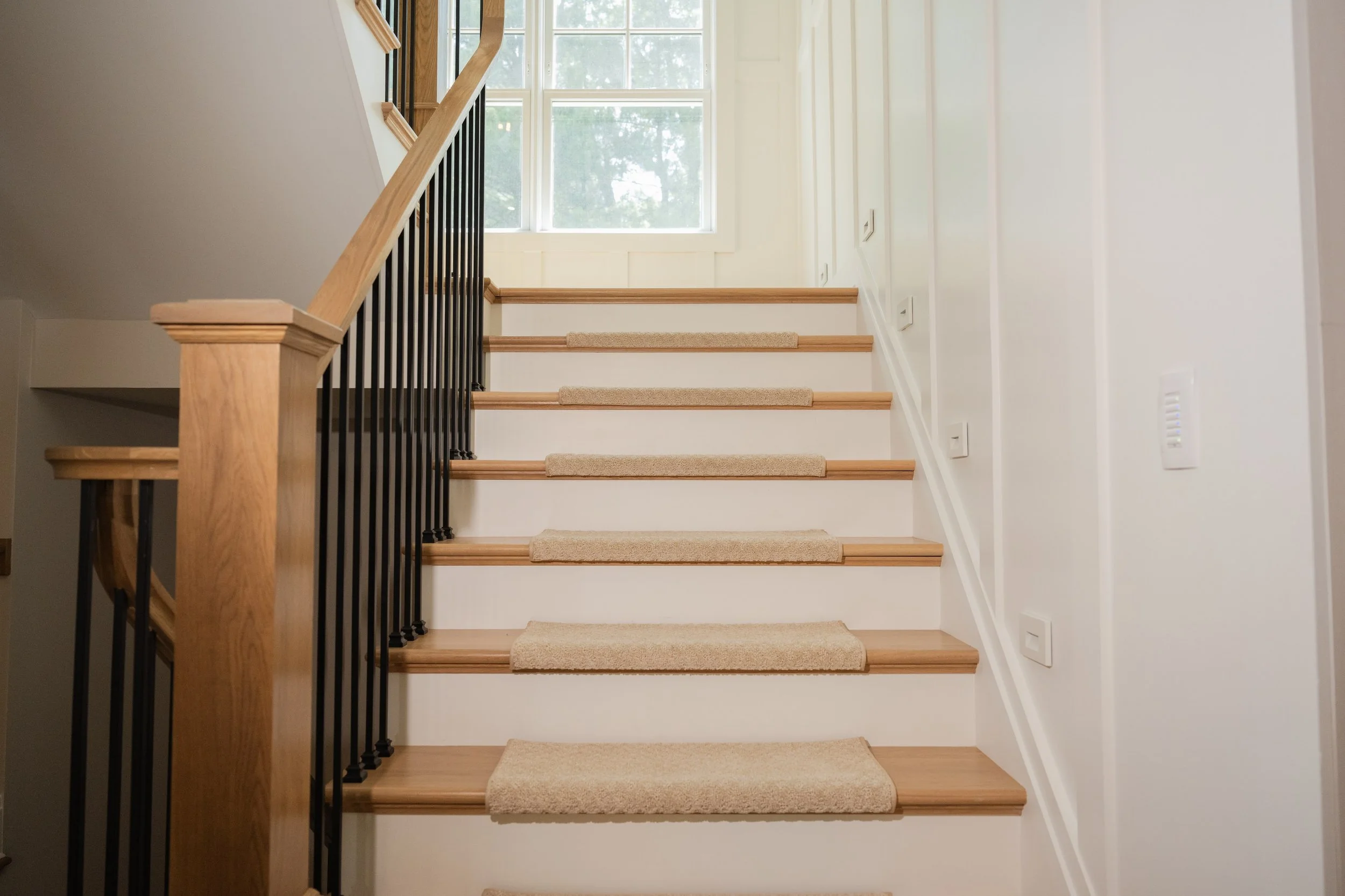 Interior view of a staircase with wooden steps and a black metal railing, leading up towards a large window letting in natural light, with white walls and beige carpet on the steps.