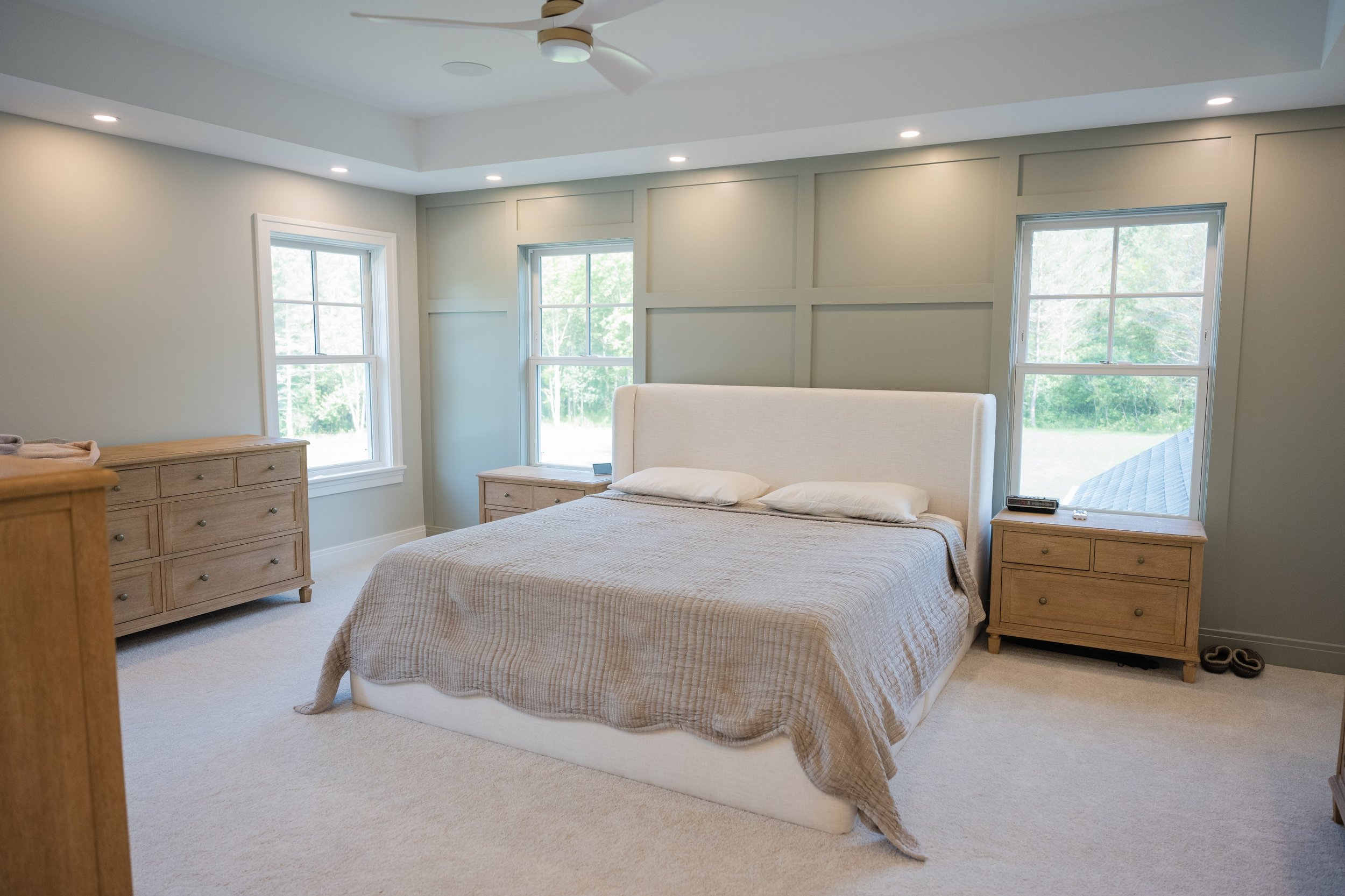 A bedroom with a white bed, beige bedding, and two wooden nightstands. There are three large windows with white frames and a view of trees outside. The walls are painted light green, and the ceiling has recessed lighting and a white ceiling fan.