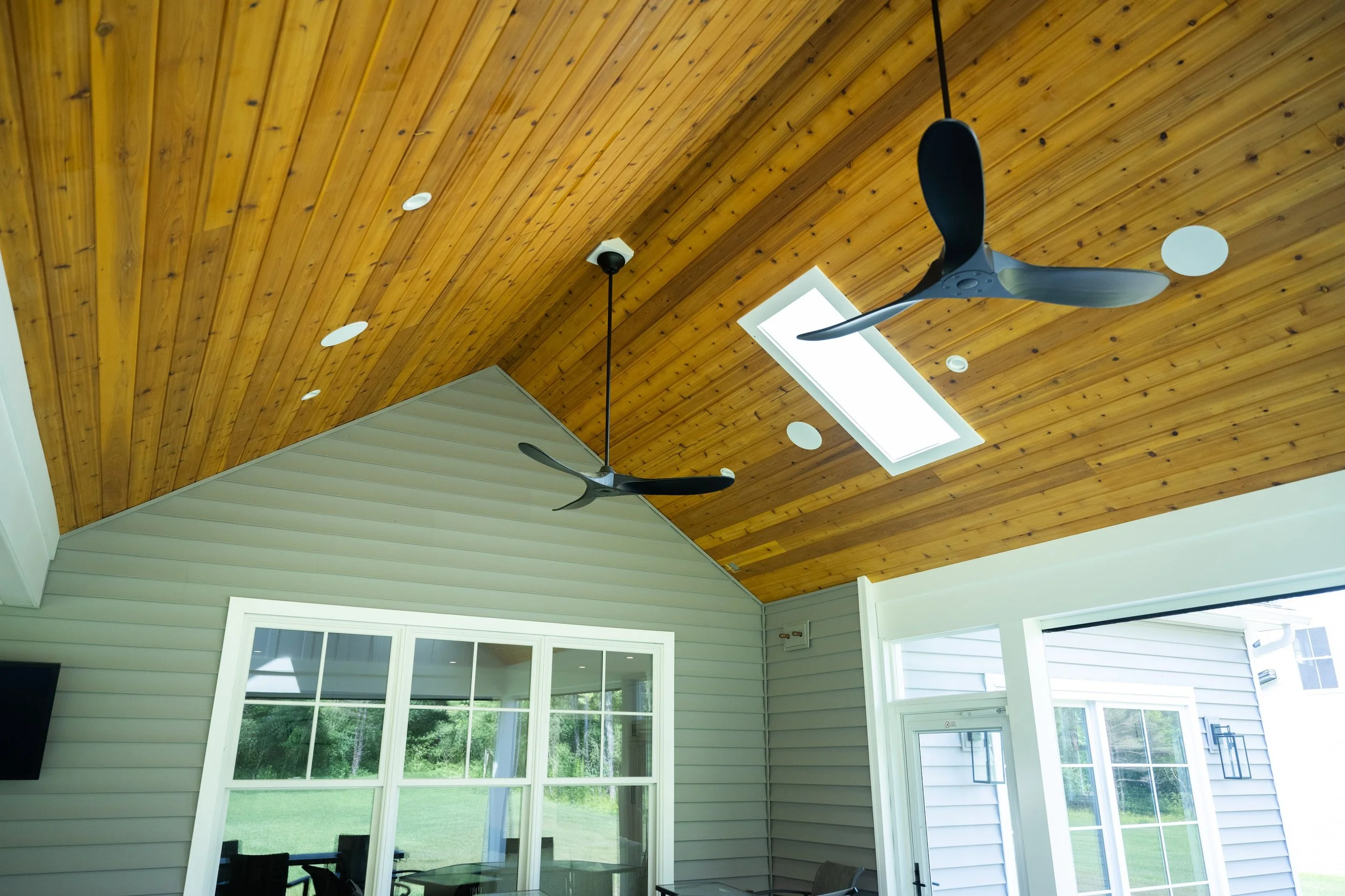 Interior view of a room with a wooden ceiling, two black ceiling fans, a skylight, and large windows showing greenery outside.