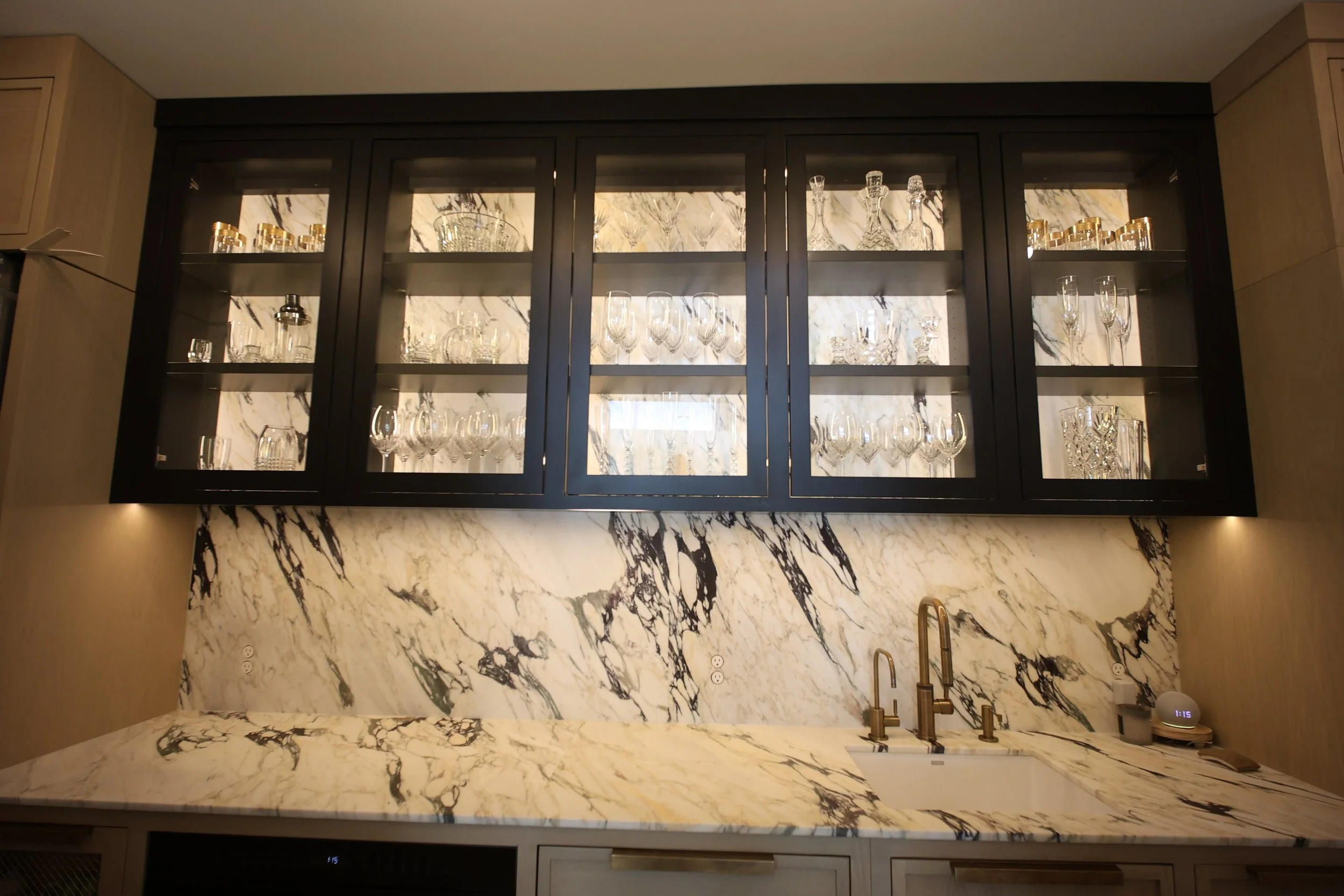 A modern kitchen featuring a marble countertop and backsplash with black and white veins, a black upper cabinet with glass doors displaying glassware, a gold faucet, and a small digital clock with blue display in the corner.