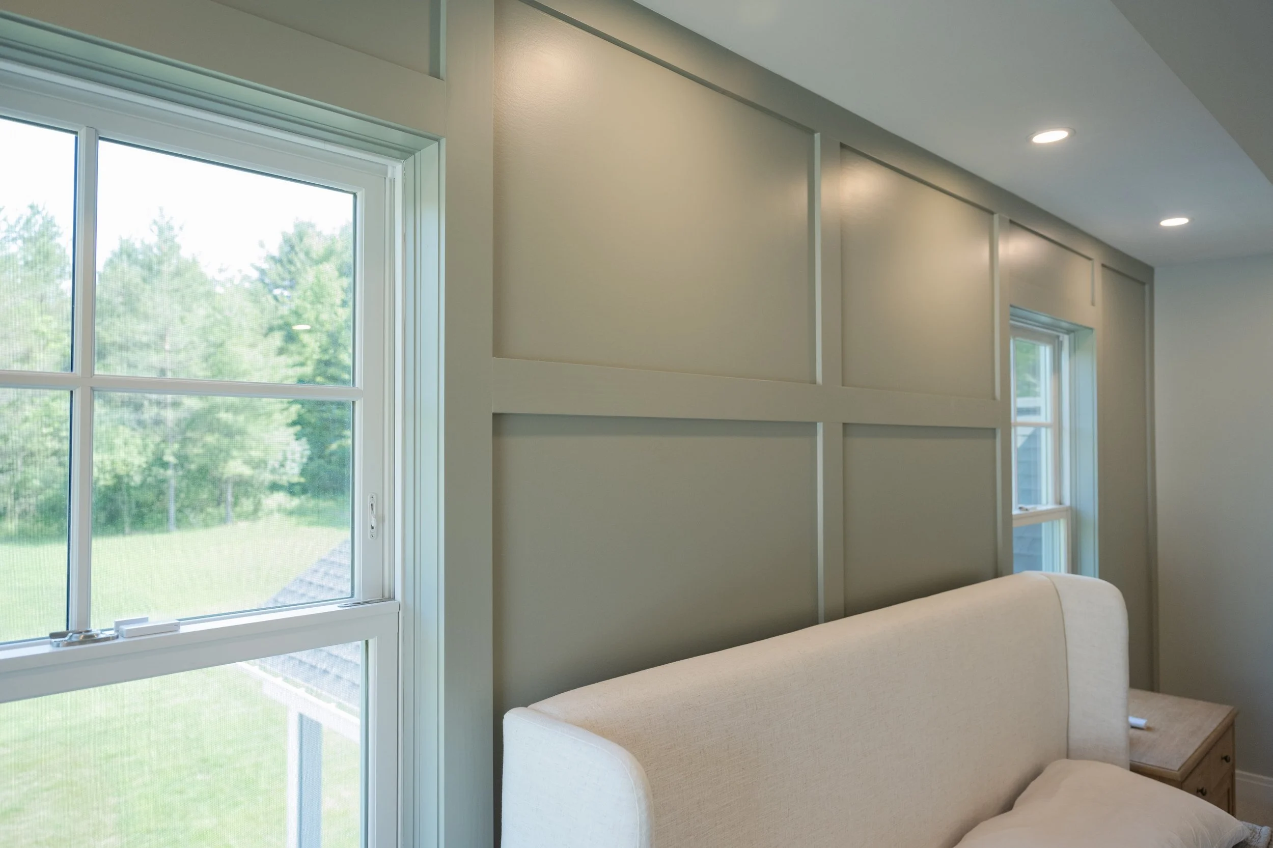 The image shows a corner of a room with large windows letting in natural light. The walls are painted light gray, and there are built-in wall panels. Two recessed ceiling lights are visible, and a beige armchair with part of a wooden side table is in