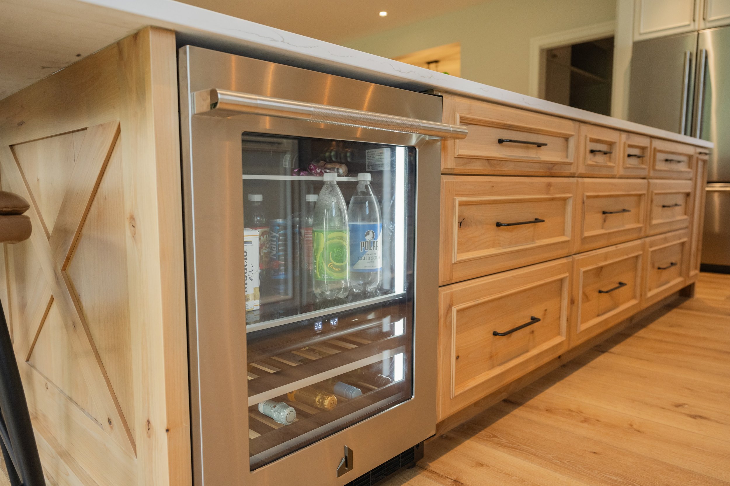 A wooden kitchen counter with black handles, and a small glass-door refrigerator containing bottled drinks. There are various bottles and cans inside the fridge, and the floor is wood.