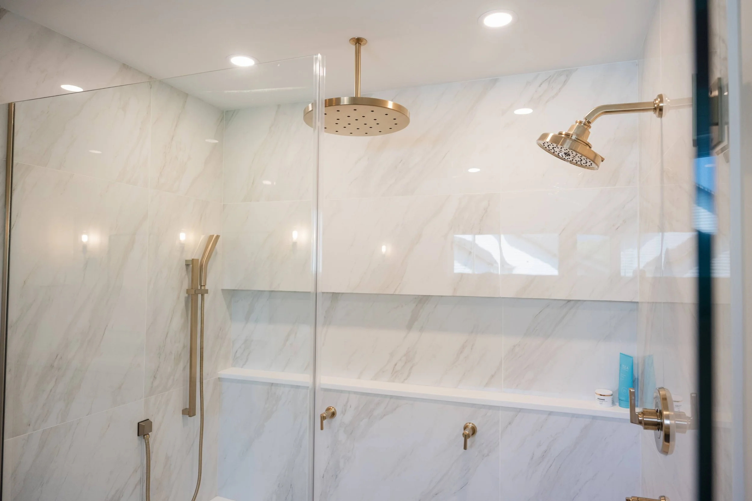 A modern shower area with beige marble tiles on the walls and ceiling. It features a large round ceiling-mounted rainfall showerhead and a smaller wall-mounted showerhead, both in a gold-colored finish. There is a built-in shelf with personal care pr