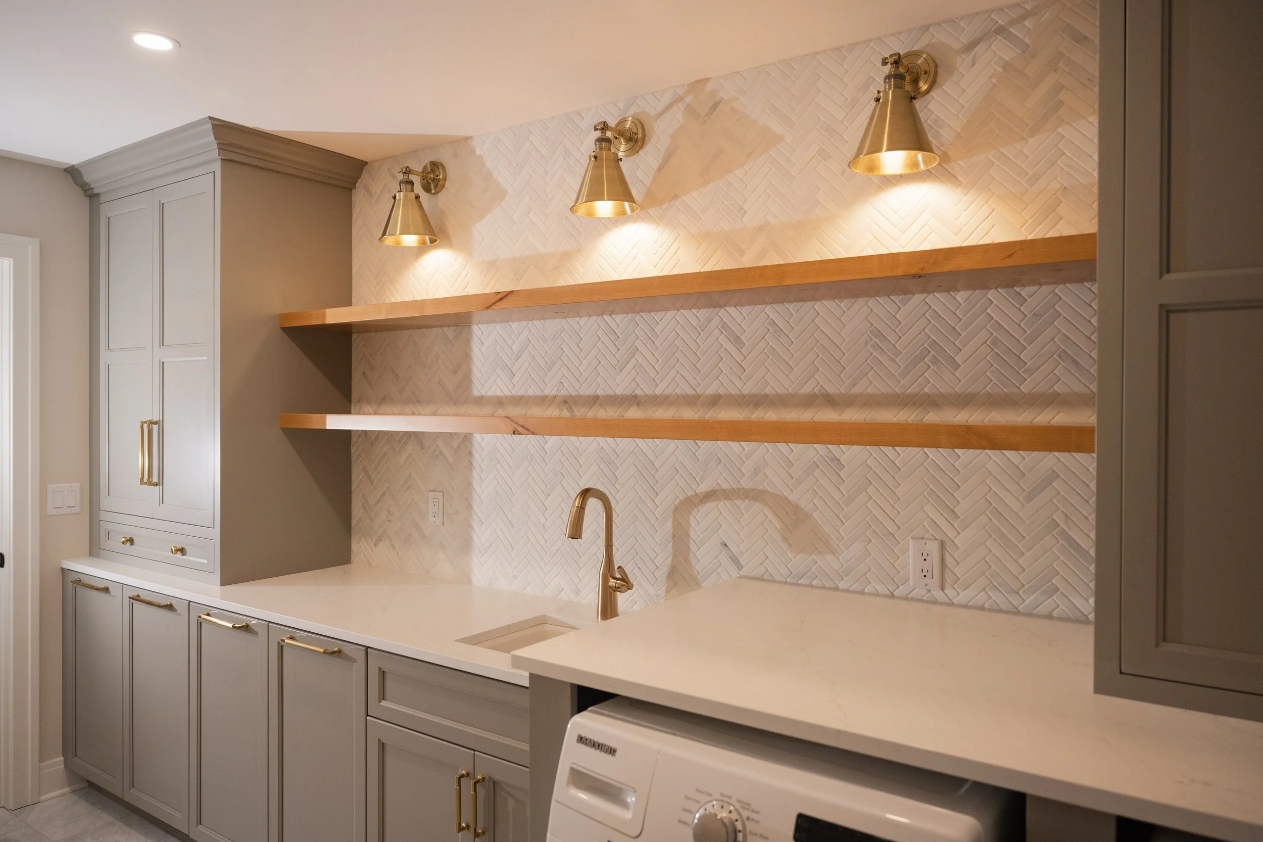 A modern kitchen with off-white cabinetry, a white countertop, and a decorative backsplash with a herringbone tile pattern. Three brass wall-mounted lamps illuminate the backsplash area. There is a small sink with a brass faucet and a washing machine