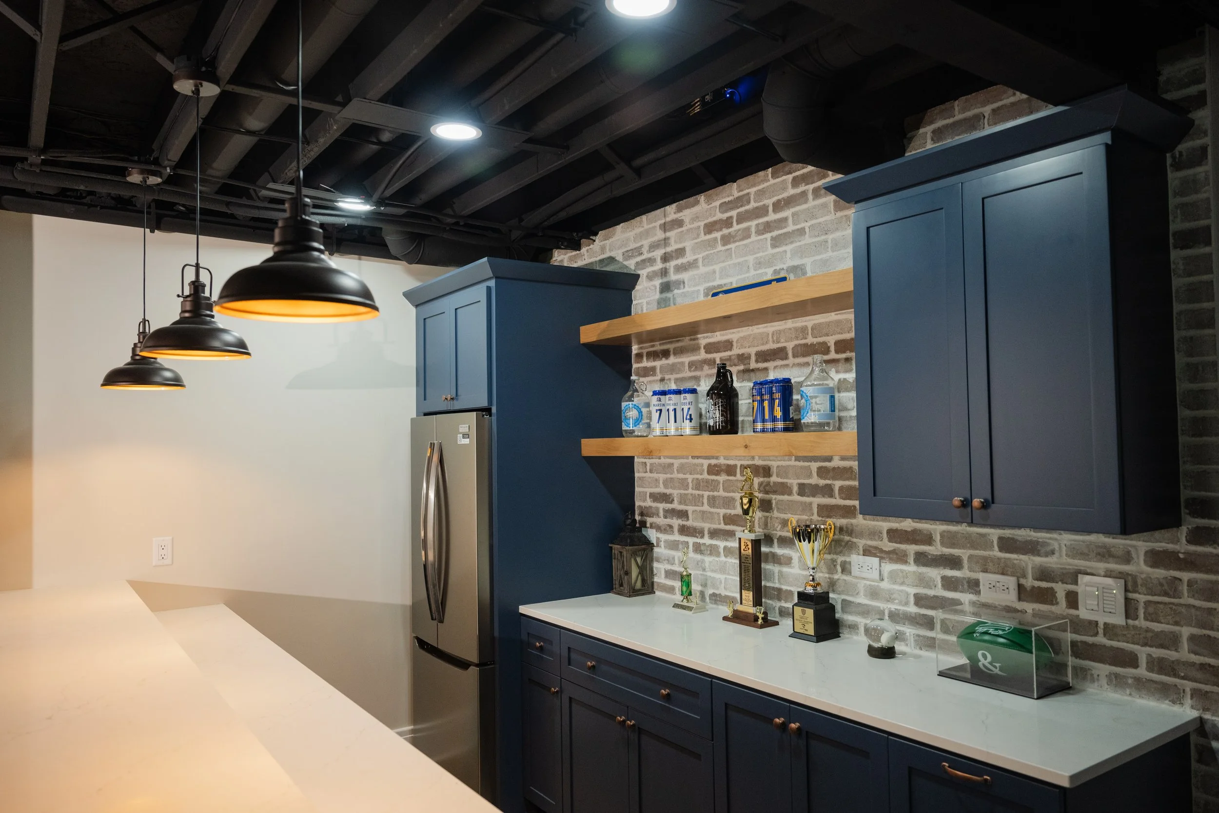 A modern kitchen with navy blue cabinetry, a light-colored countertop, and a brick accent wall. Three black pendant lights hang from the ceiling, which has exposed black beams and ductwork. The kitchen displays several trophies and awards on a shelf 