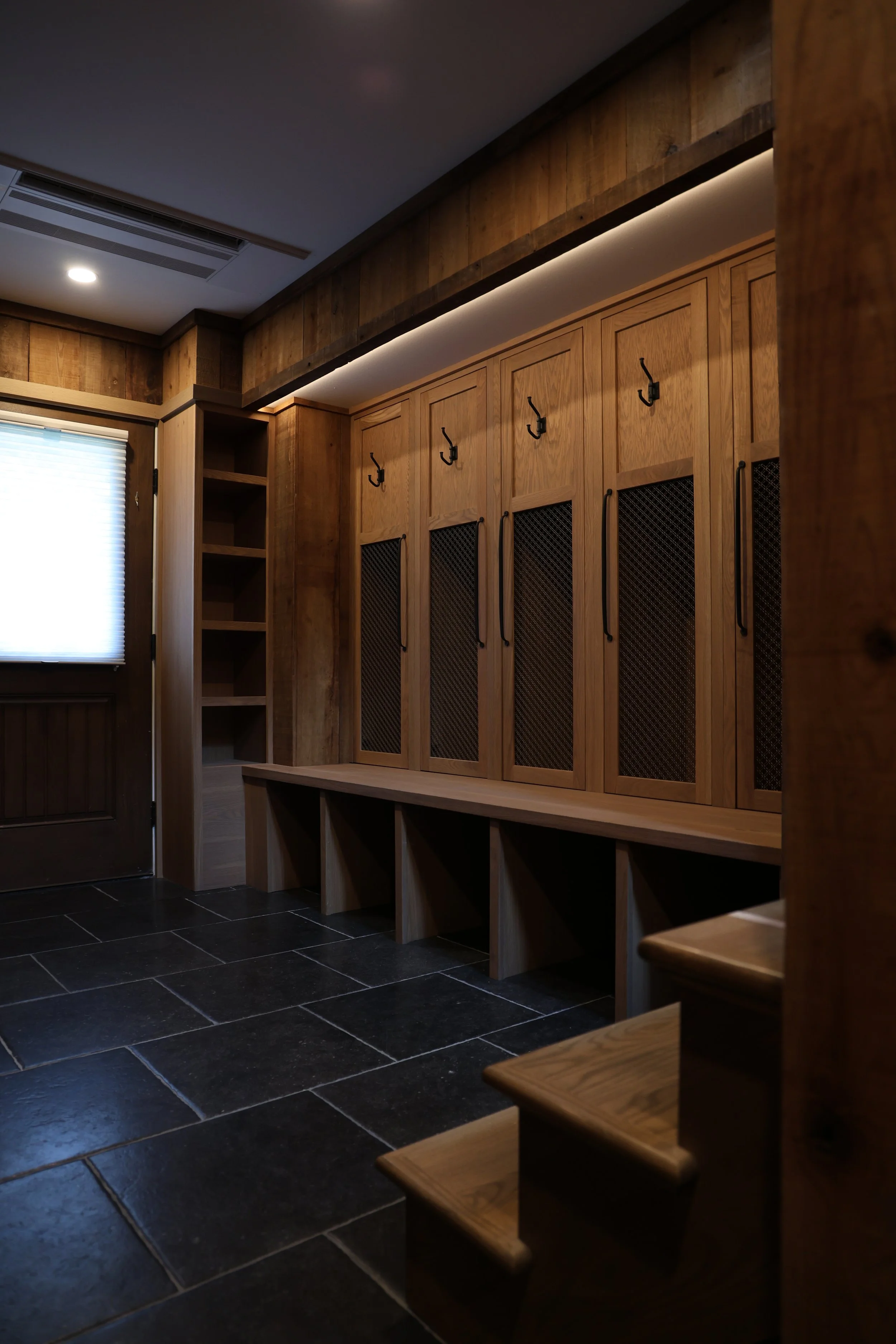 Wooden lockers with black hooks in a room with dark tiled floor and a window with closed blinds.