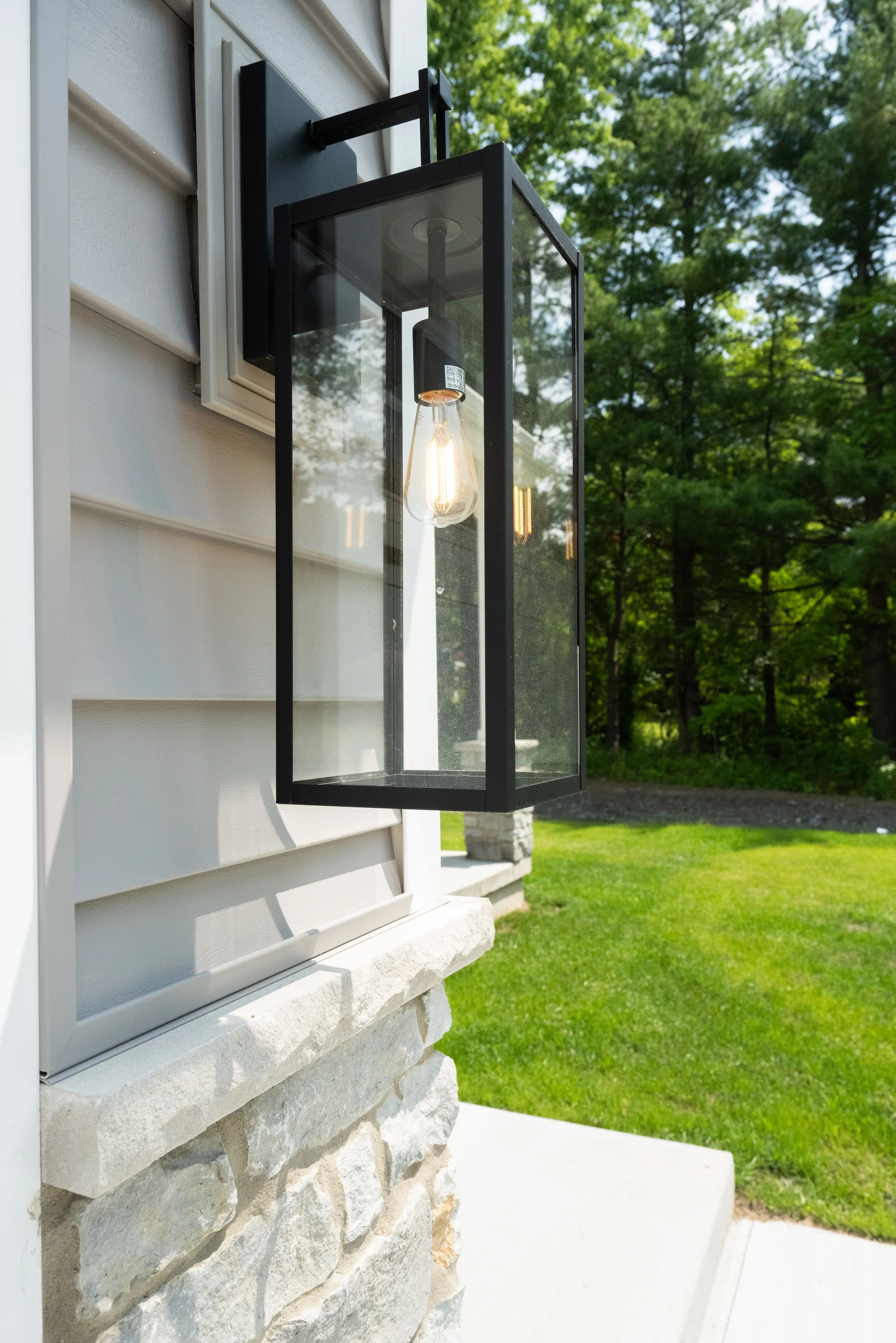 Close-up of a black outdoor lantern light fixture mounted on the side of a house with beige siding, with trees and a lawn in the background.