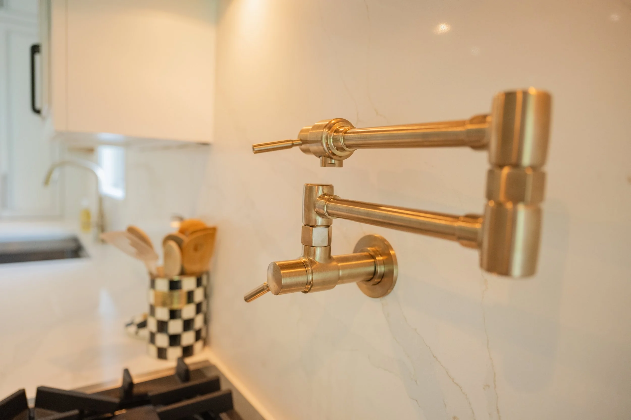 Close-up of a modern brass kitchen faucet mounted on a white marble countertop in a kitchen. In the background, a checkered black and white cup and a white cabinet are visible.