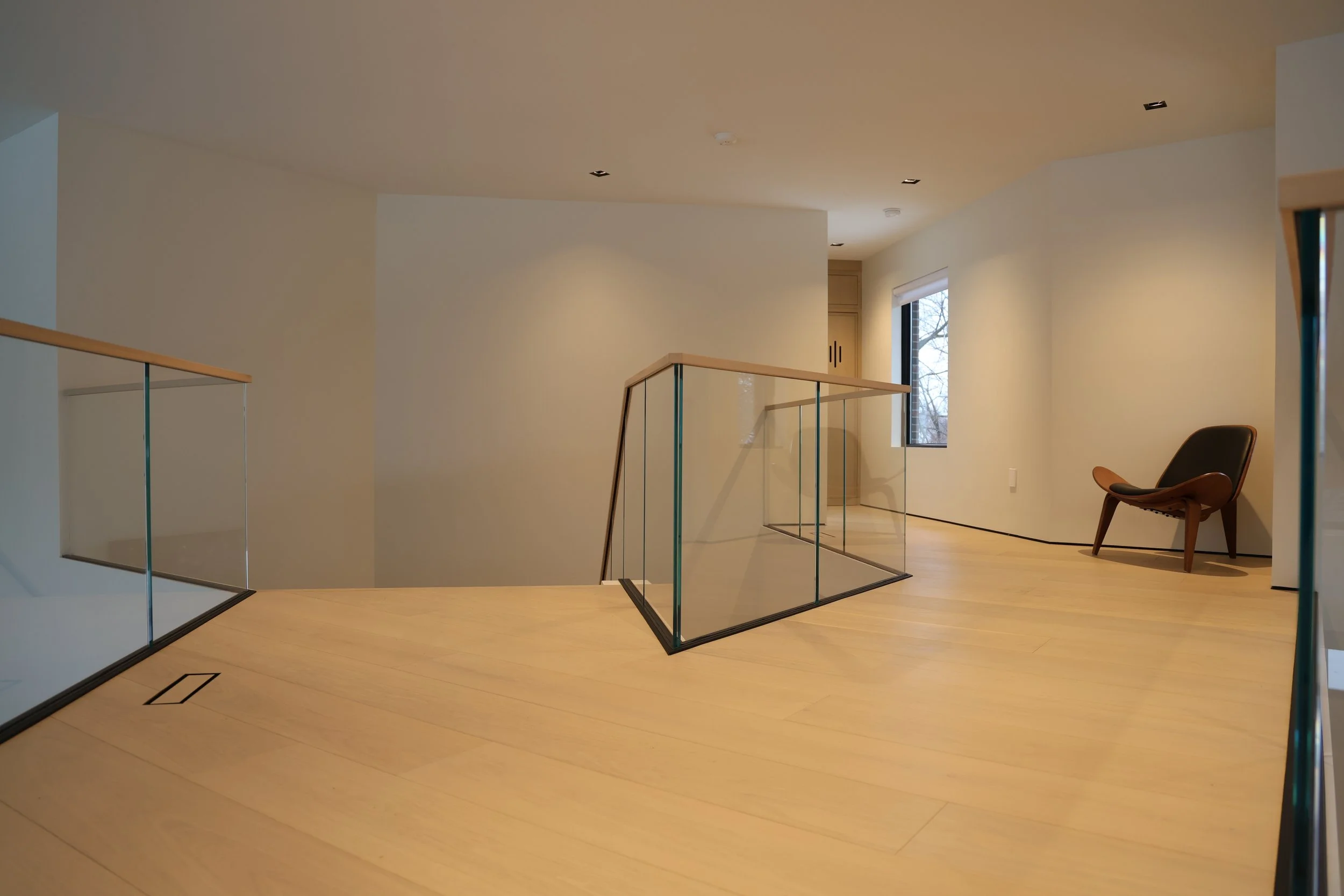 Interior view of a modern, minimalist living space with wooden floors, white walls, a window, and a black chair. There is a staircase with glass railings and a side table near the window.