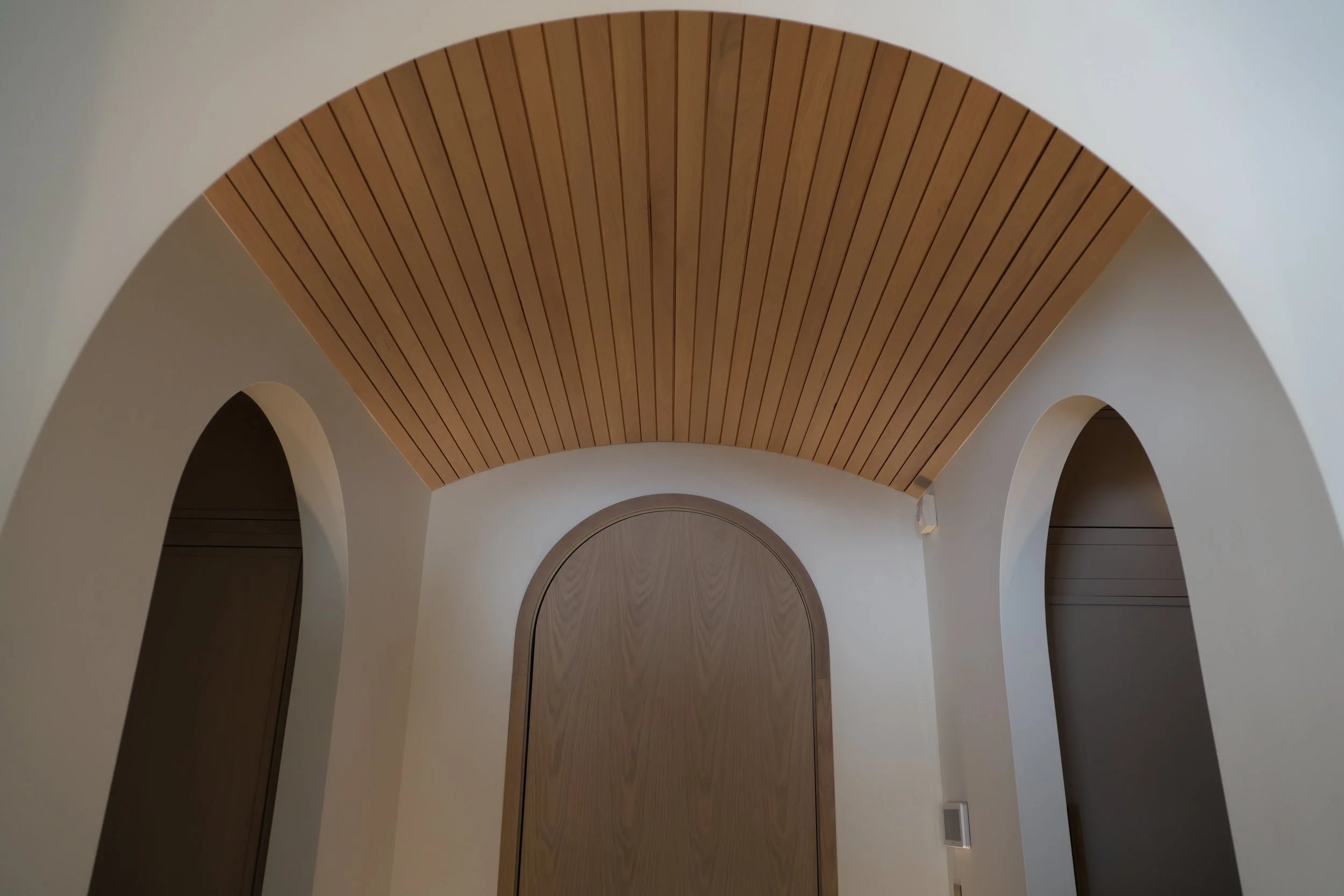 View of a ceiling with wooden slats arranged in parallel lines, framed by a white curved archway in a modern interior space.