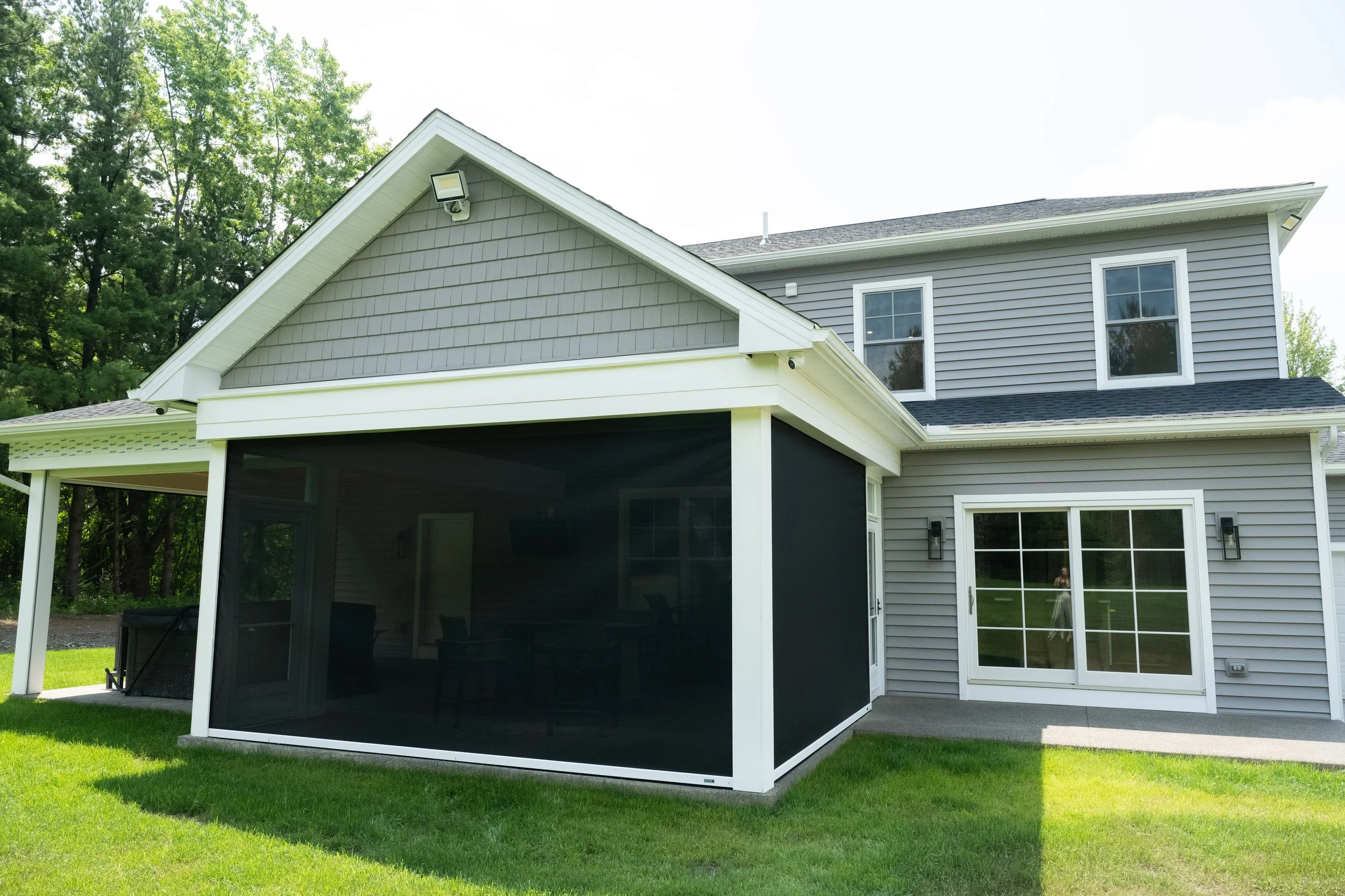 Backyard view of a two-story house with gray siding, multiple windows, and a screened-in porch with black mesh. There's a lush green lawn and trees in the background under a bright sky.